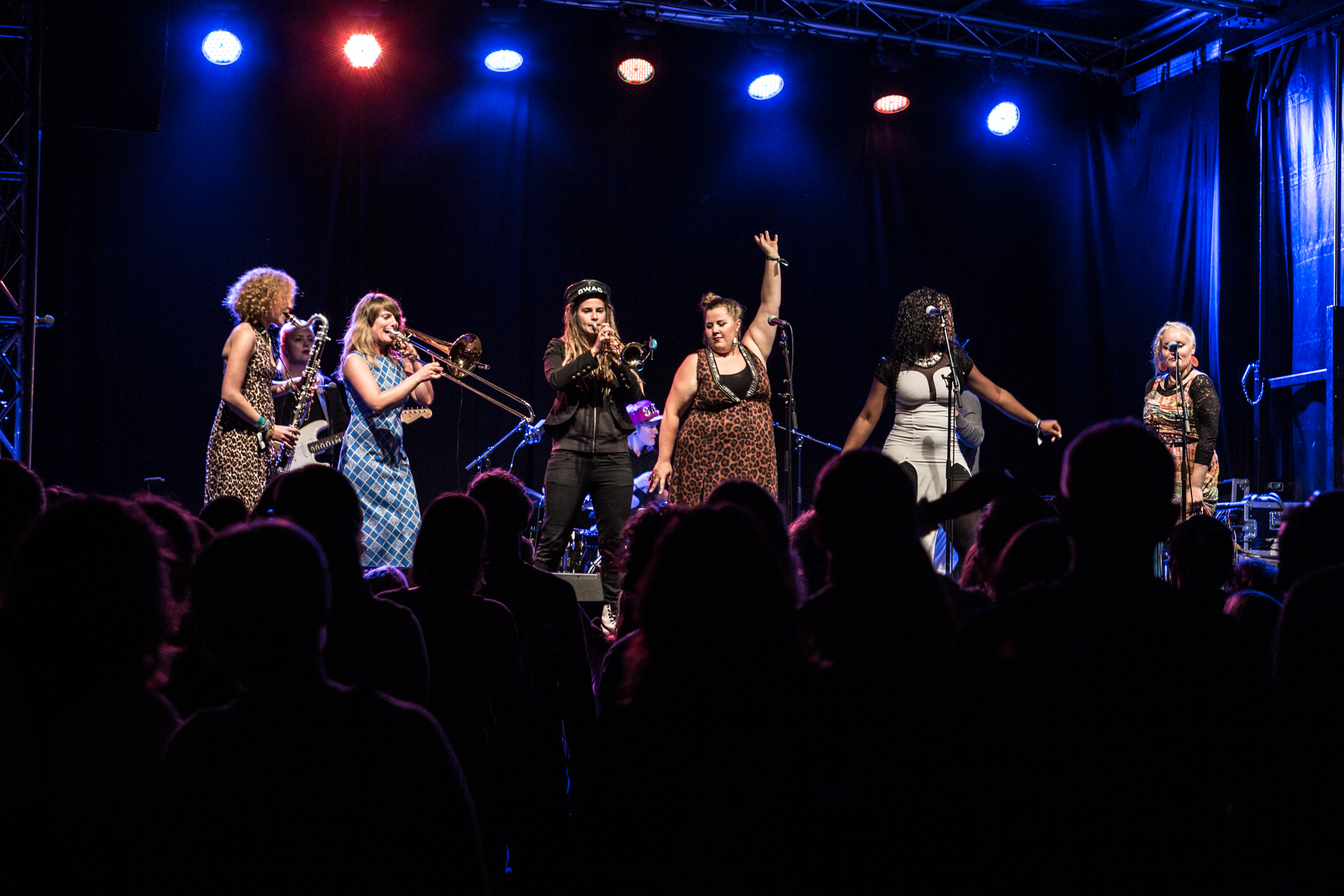 Sju kvinnligt kodade personer uppträder på en scen med publik framför.