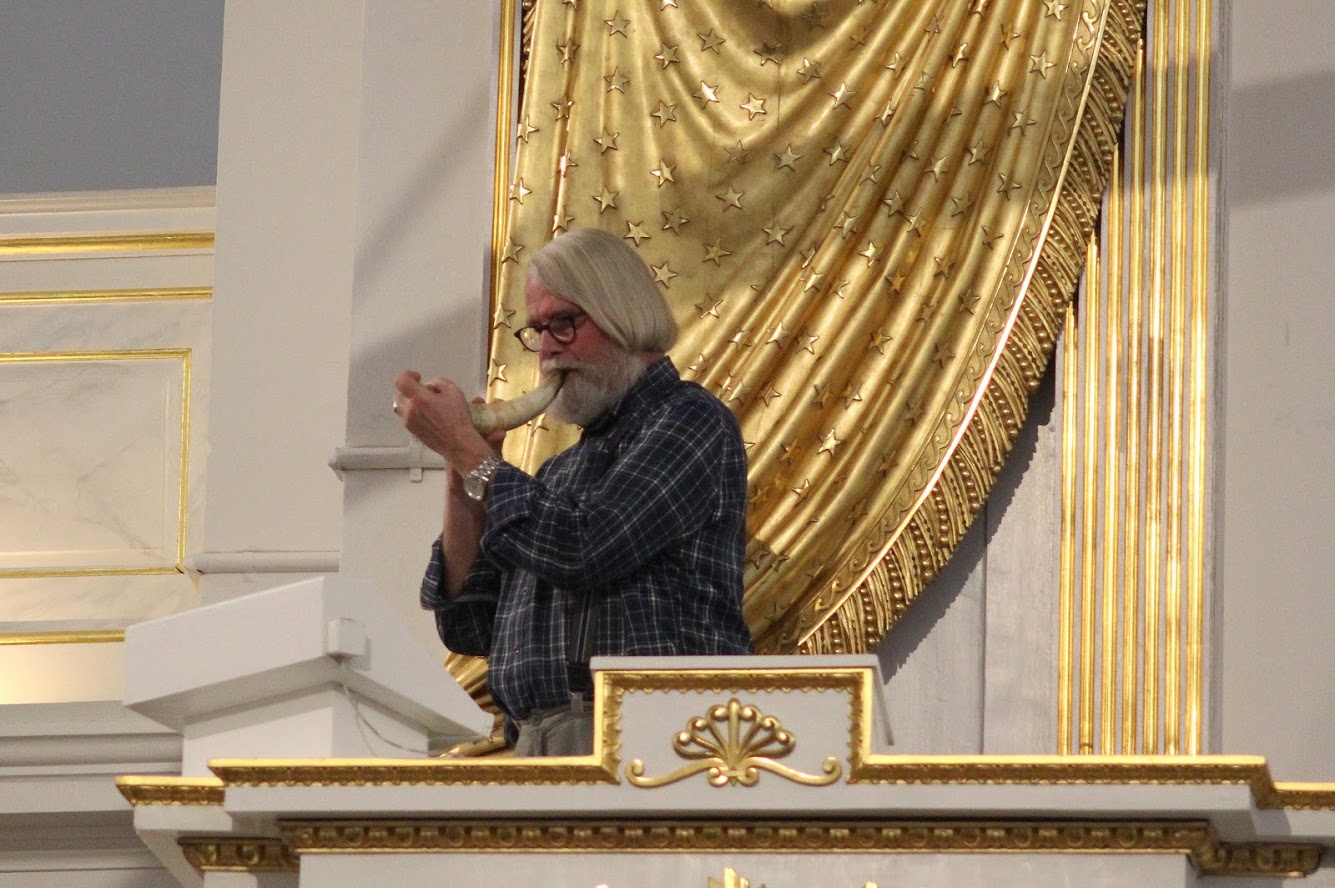 A male coded person with white hair, glasses and checkered shirt plays in a horn. They stand in a pulpit in a church with a golden veil in the background.