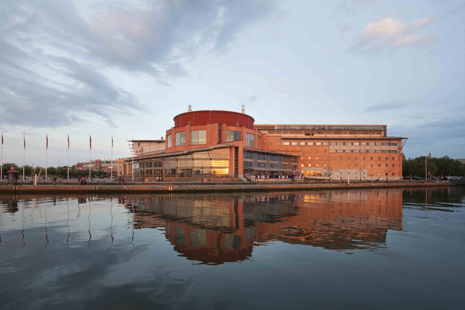 GöteborgsOperan en sommarkväll. Byggnaden är i rött tegel och med stora glaspartier. Framför byggnaden finns en brygga och därfter kommer Göta Älv.