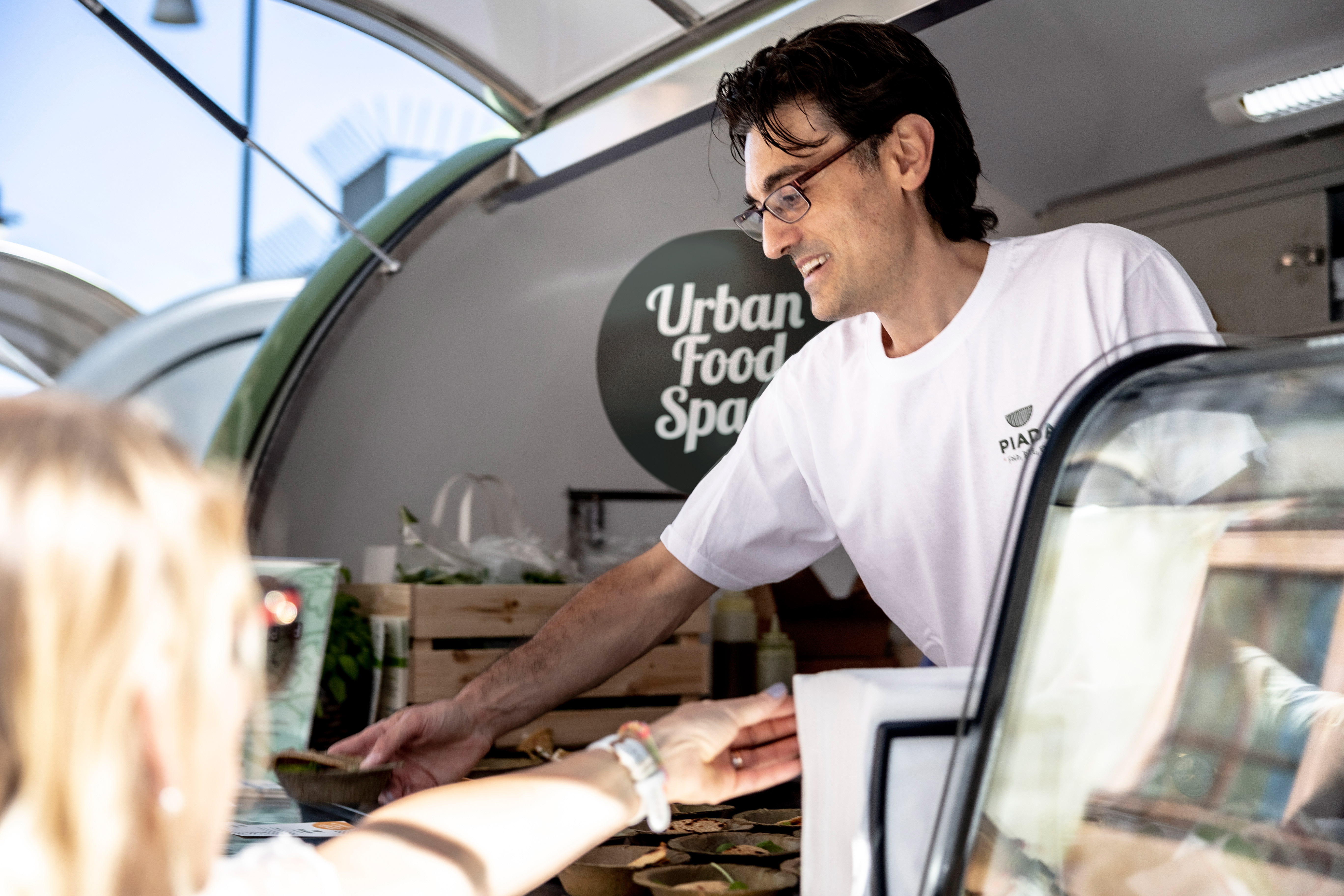 A male coded person in a white t-shirt stands in a food truck serving guests.  Inside the food truck the Urban Food Space logotype is visible.