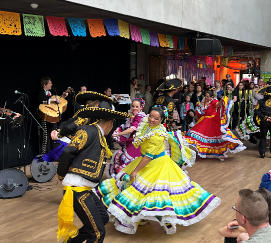 Personer i traditionella mexikanska kläder dansar. I bakgrunden spelar ett mexikanst band.