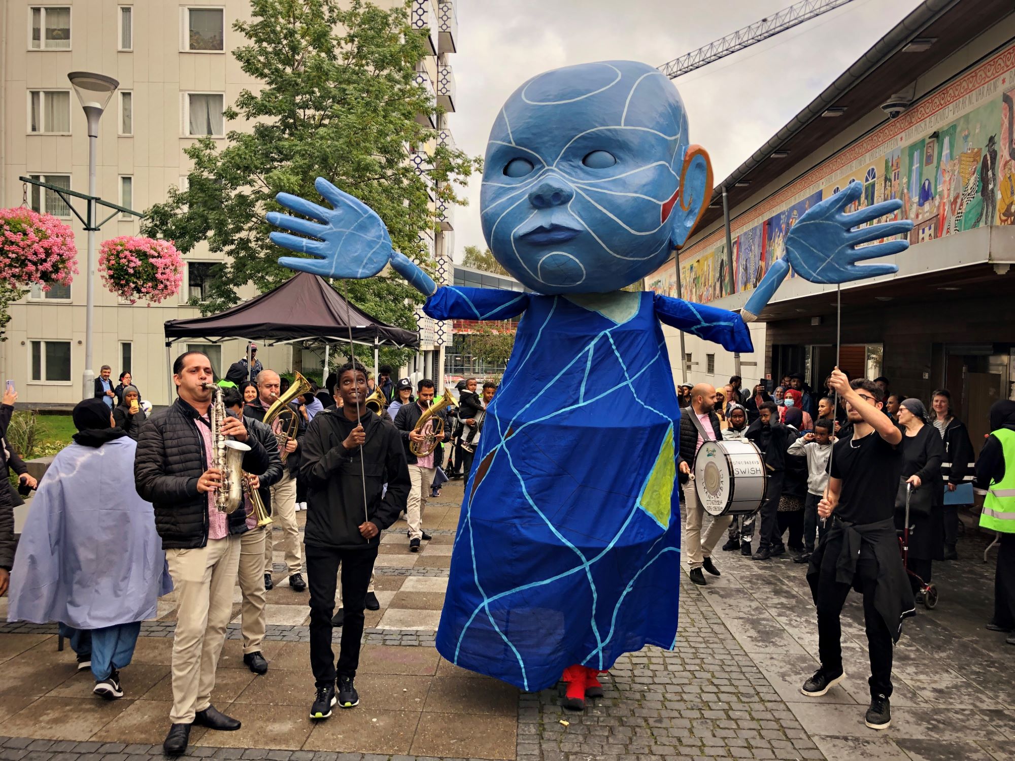 En blå figur med ett stort huvud syns gå längs med Kulturstråket i Bergsjön. Är det den festliga figurparaden? Människorna som betraktar runt om vittnar om att det verkar vara så.