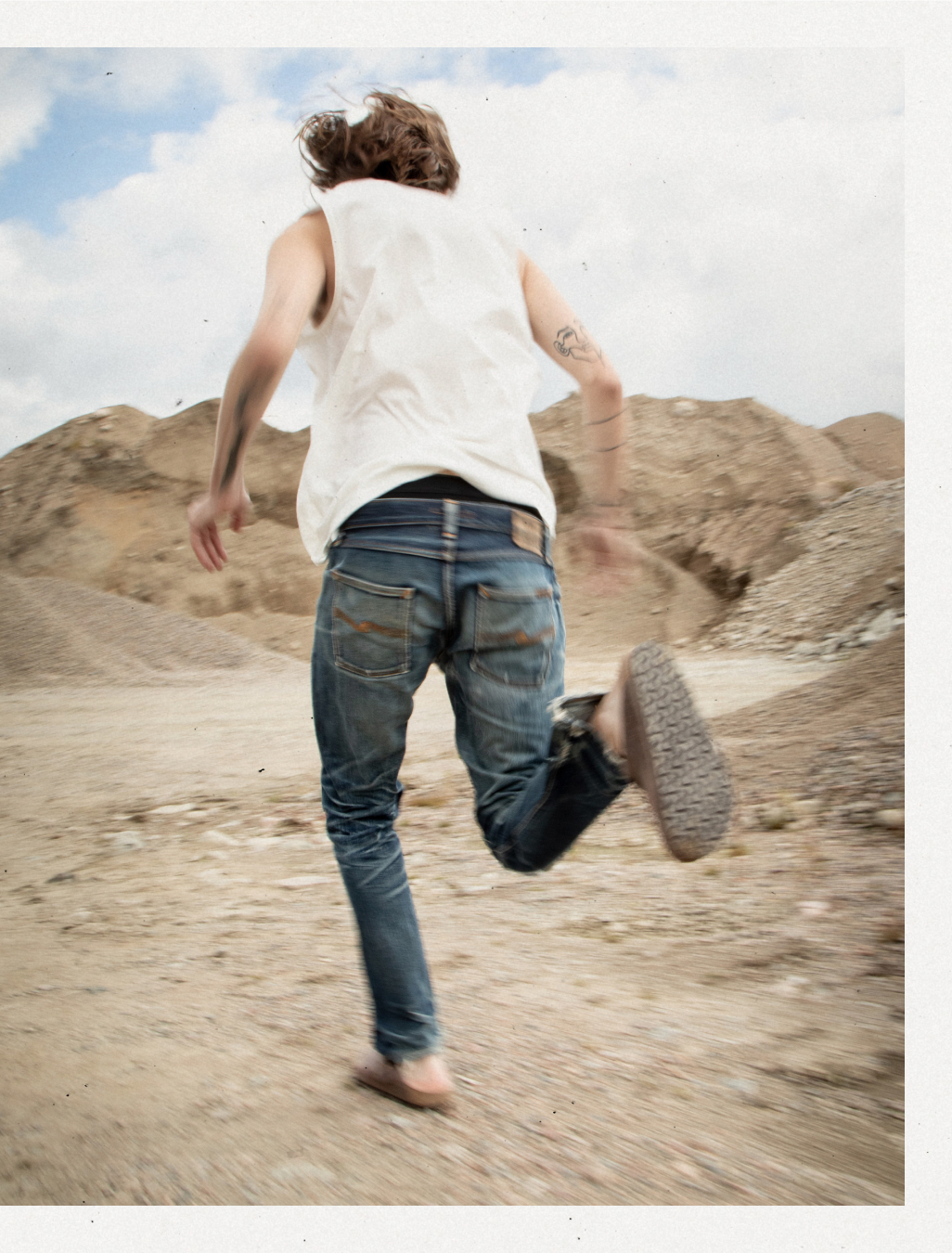 En person med jeans, vit t-shirt och tofflor syns springa från kameran. Omgivningen är fylld med grus/sand-högar. blå himmel och moln.