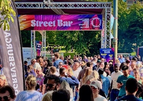 En färgstark folkmassa rör sig under ett skyltat entrétak med texten “Street Bar” i stora bokstäver.