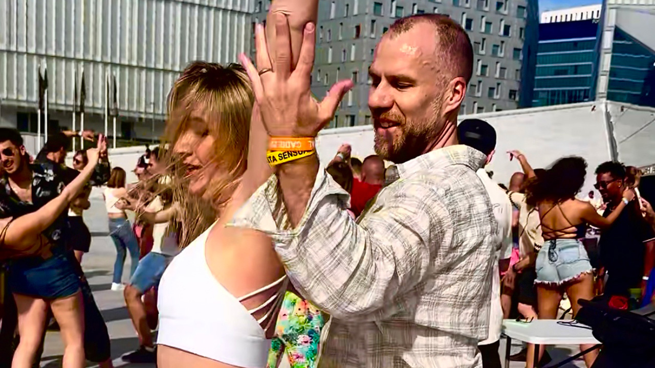 En man och en kvinna dansar tillsammans utomhus bland en grupp människor. Båda ser glada ut och omgivningen är fylld av rörelse och musik.