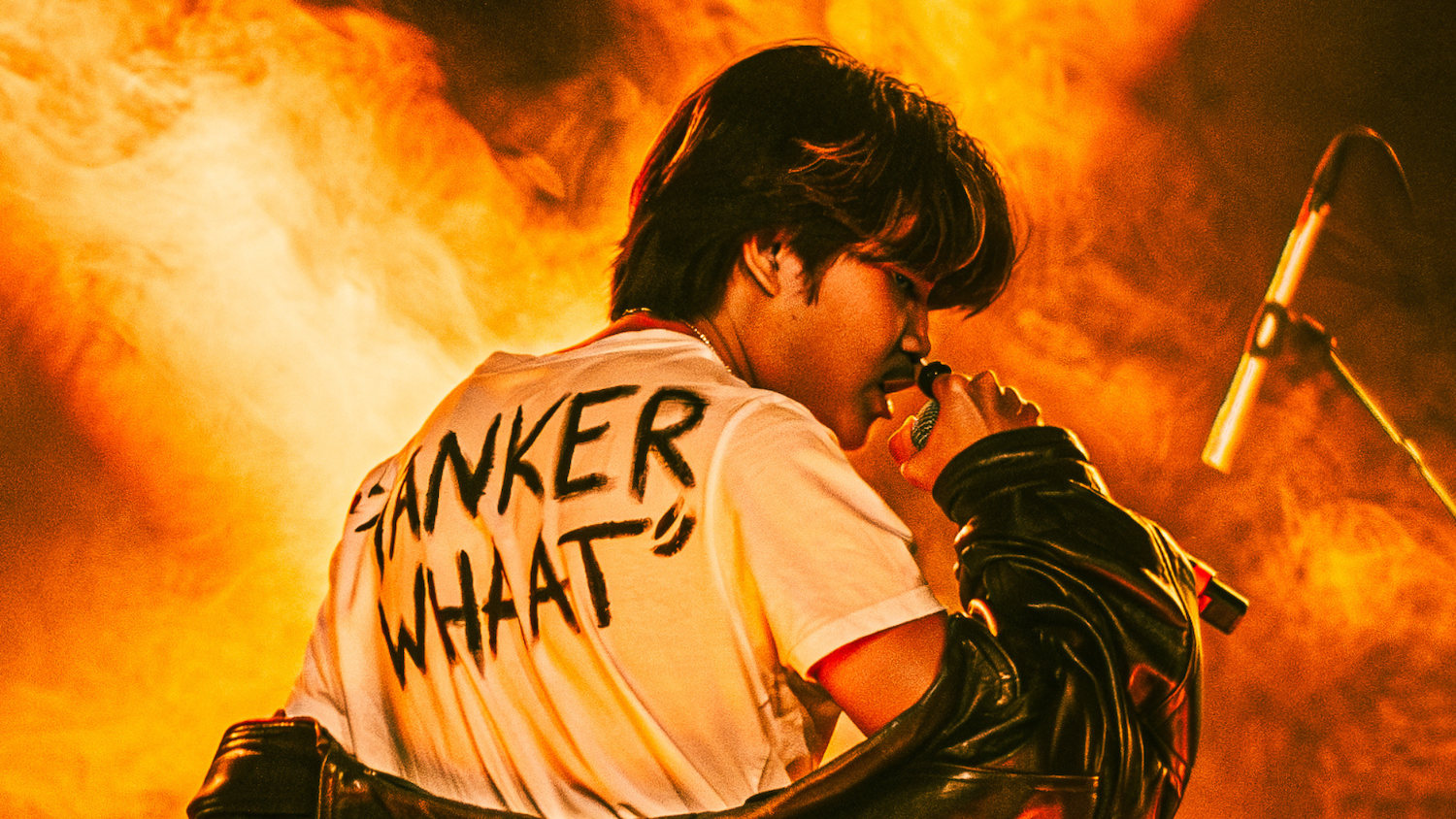 En person står med ryggen mot kameran och sjunger in i en mick. På ryggen på hans T-shirt står en svart text. Bakgrunden är orange och liknar eld.