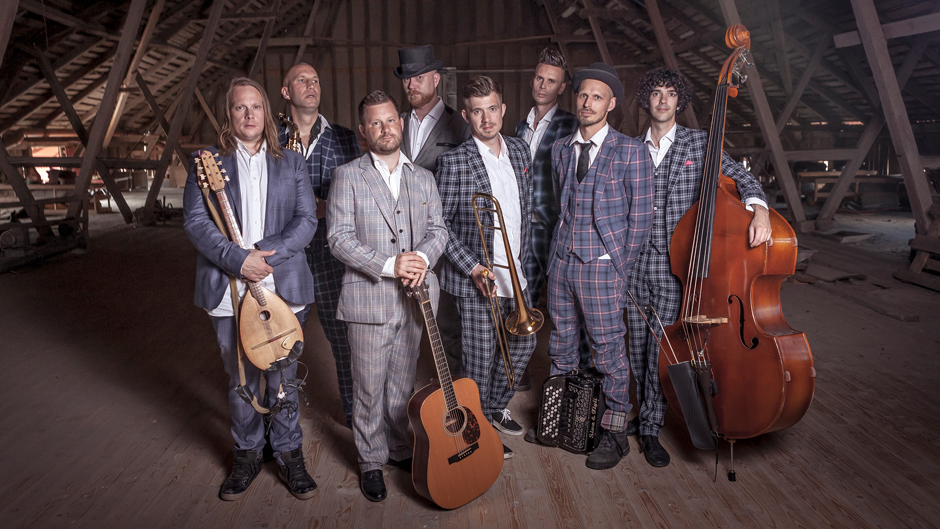 En grupp av åtta män iklädda kostym står med olika stränginstrument i händerna. På trägolv i ett vindutrymme med snetak.
