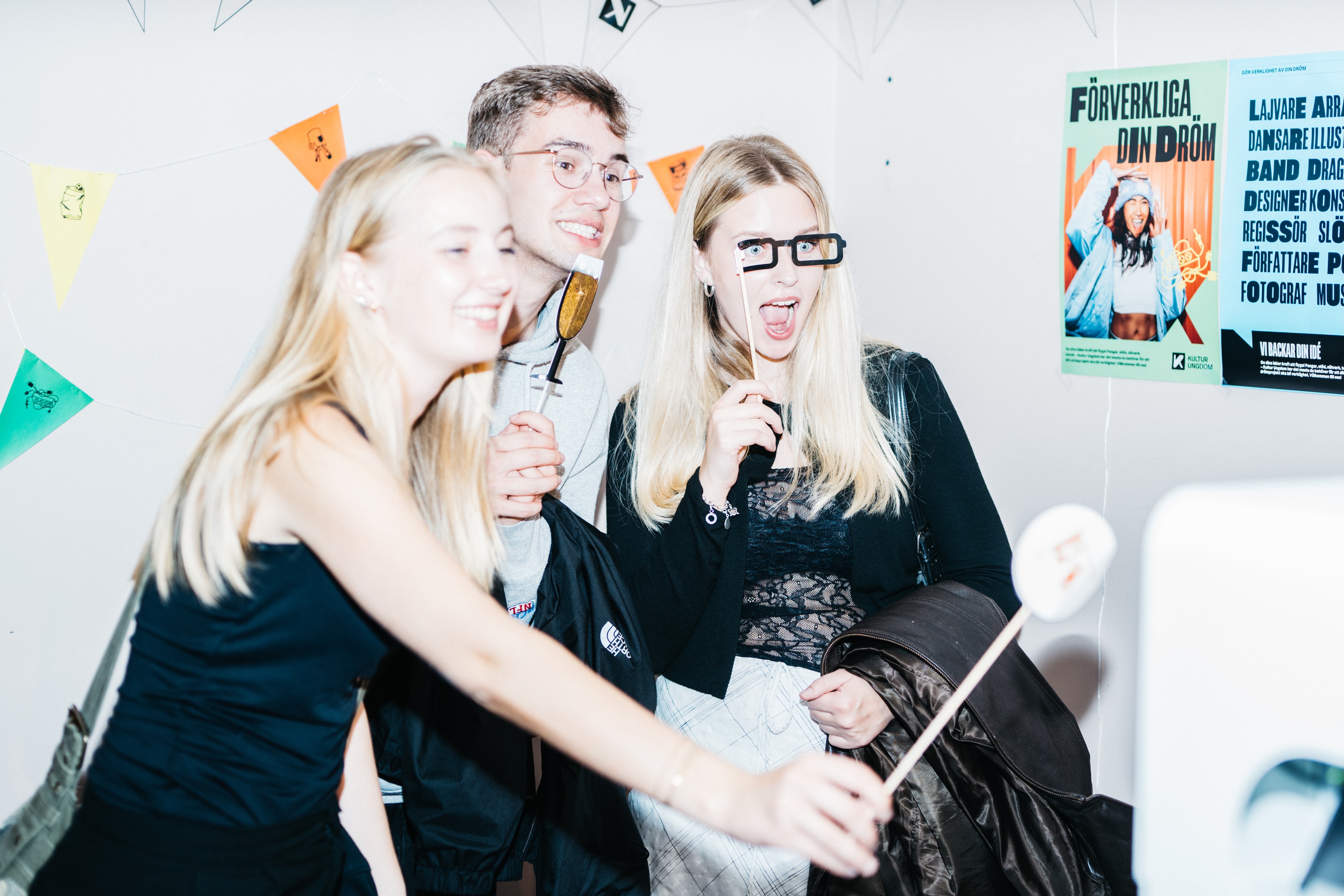Unga personer står och ler in i en kamera framför en fotovägg klädd med flaggor i olika färger.