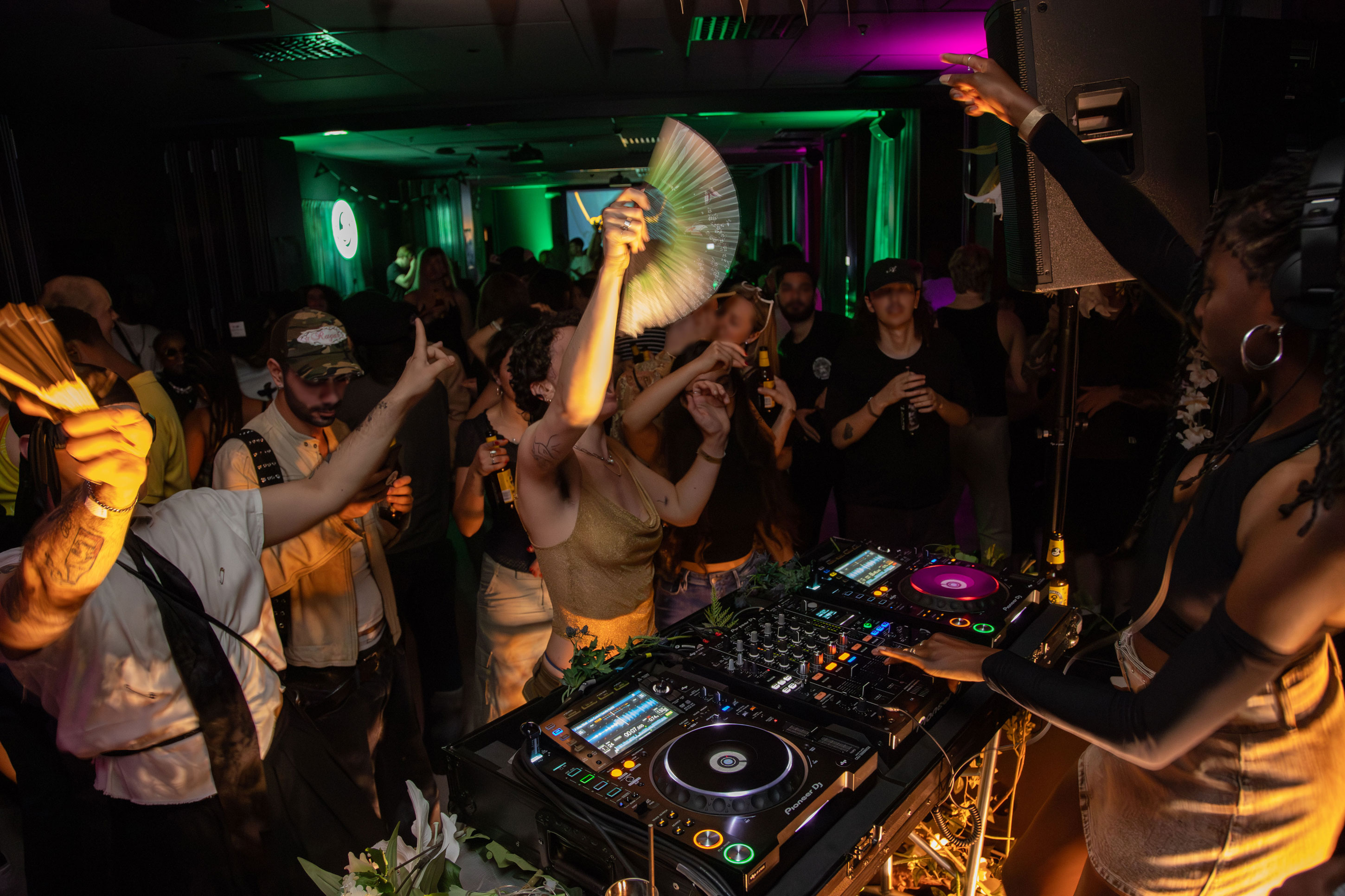 Ett dansgolv fyllt med människor som dansar och håller upp solfjädrar. Ljuset är färgglatt med gröna och lila nyanser. Till höger står en DJ vid ett mixerbord med hörlurar på sig, mitt i spelningen.