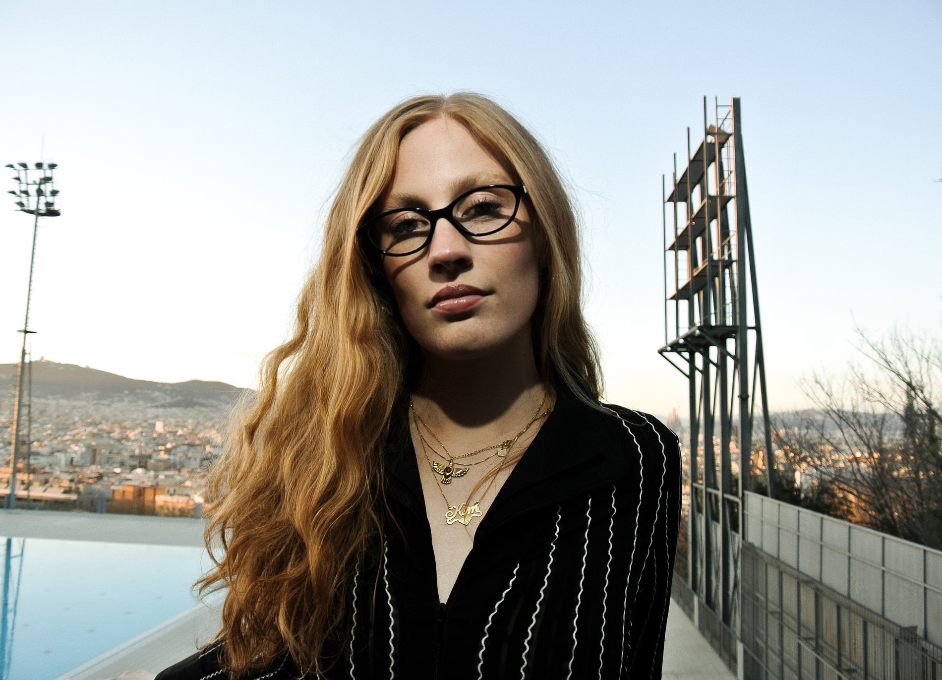 En kvinna med långt, vågigt, blont hår och glasögon står utomhus på en takterrass med utsikt över en stad och berg i bakgrunden. Hon bär en svart topp med vita ränder och flera guldfärgade halsband. Det är dagtid och himlen är klar.
