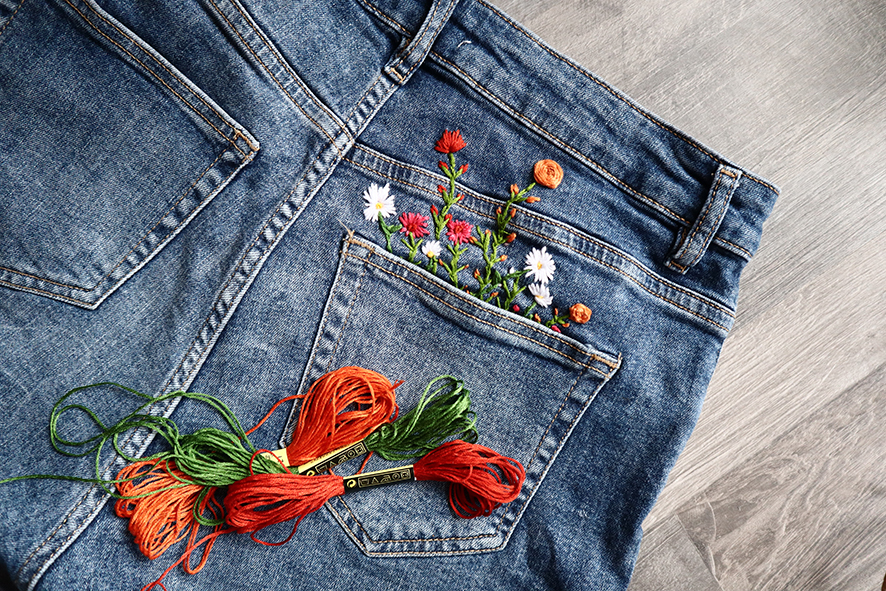 Bilden visar ett par blå jeans liggande på ett trägolv. Fokus ligger på bakfickan, där färgglada blommor är broderade så att de ser ut att växa upp ur fickan. Framför jeansen ligger flera nystan med broderigarn