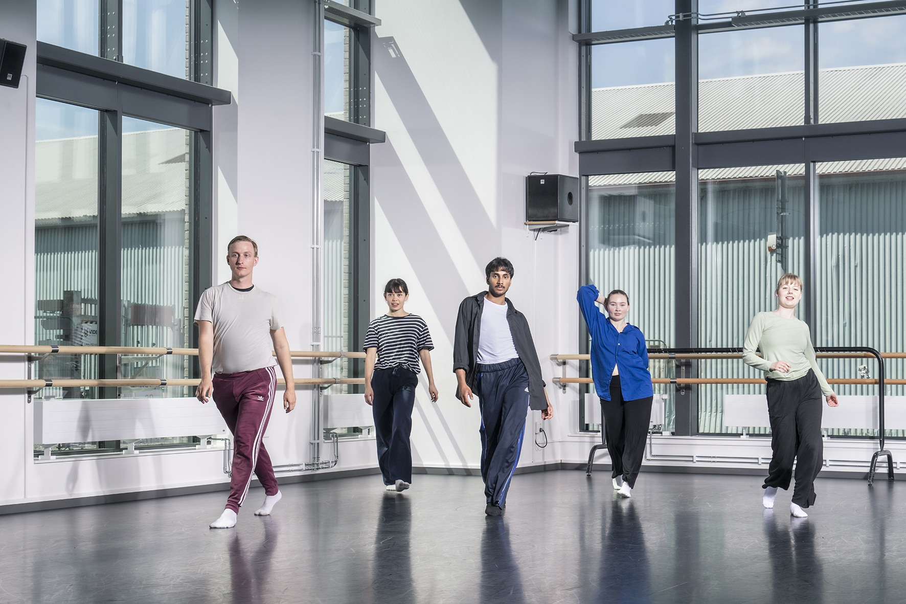 Fem dansare i ett synkroniserat danssteg. De står i en danssal med stora fönster.