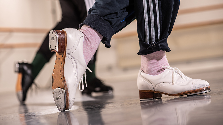 Närbild på två personer som steppar i en dansstudio. Fokus ligger på fötterna. Personen i förgrunden bär vita steppskor, rosa strumpor och mörkblå träningsbyxor med vita ränder.