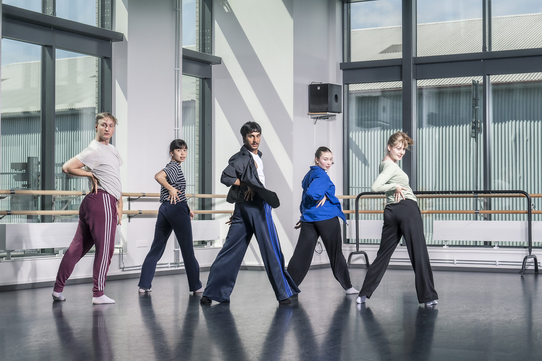 Fem dansare står i en synkroniserad danspose i en danssal med stora fönster.
