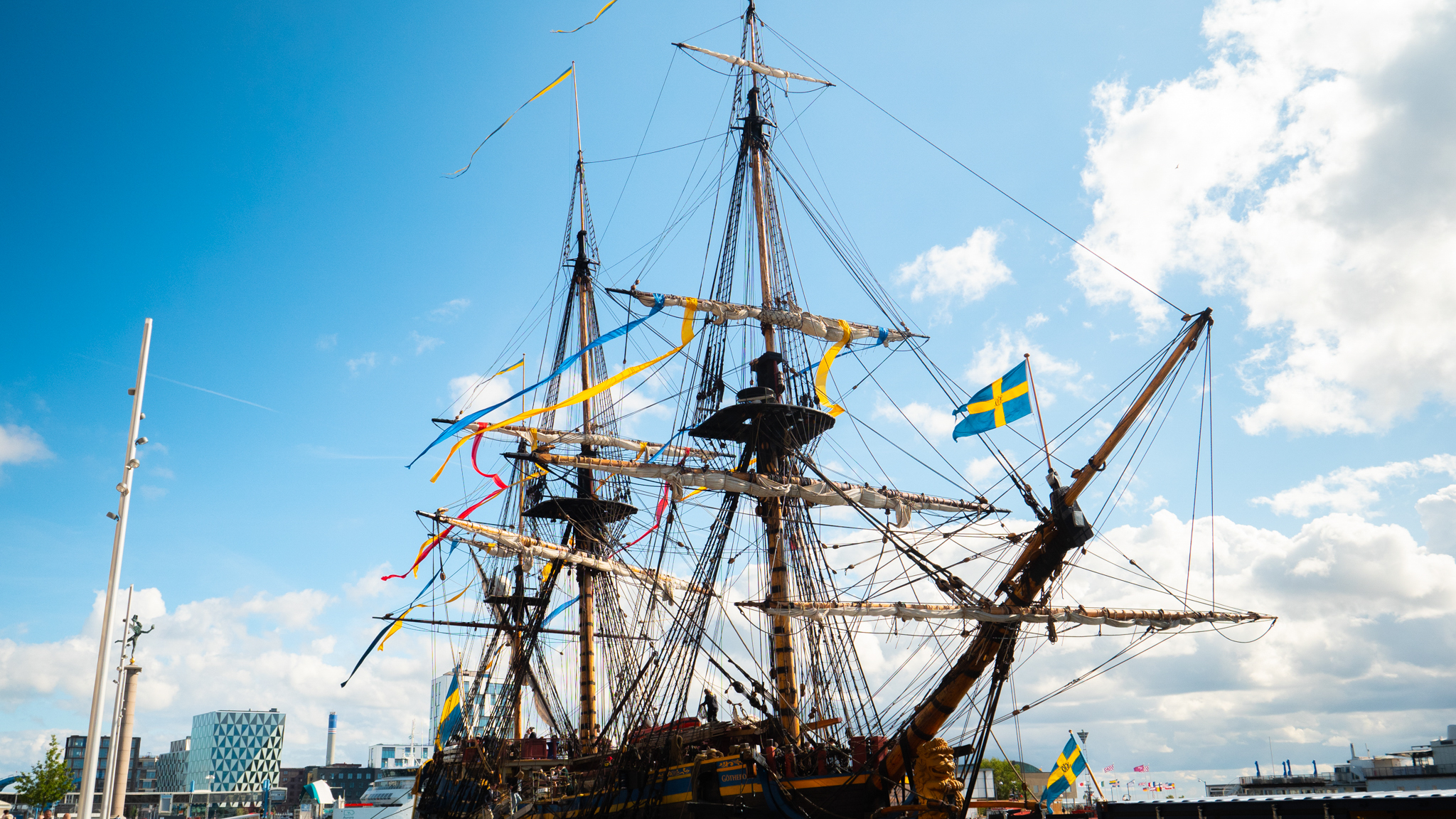 Ett stort skepp med flaggor och höga mastar. Står parkerat i en hamn med Göteborgs stadsmiljö i bakgrunden.