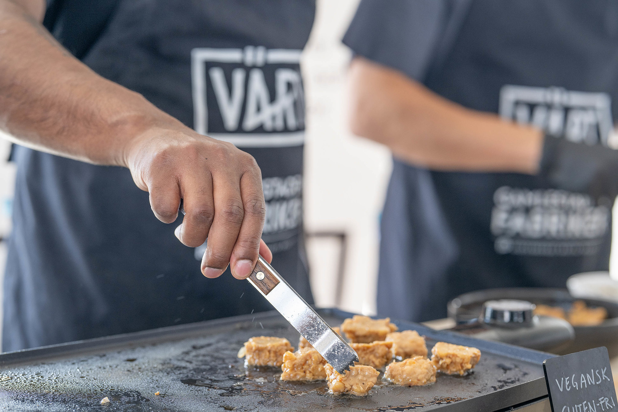 En person i förkläde använder tång för att laga vegansk mat på en stekhäll.