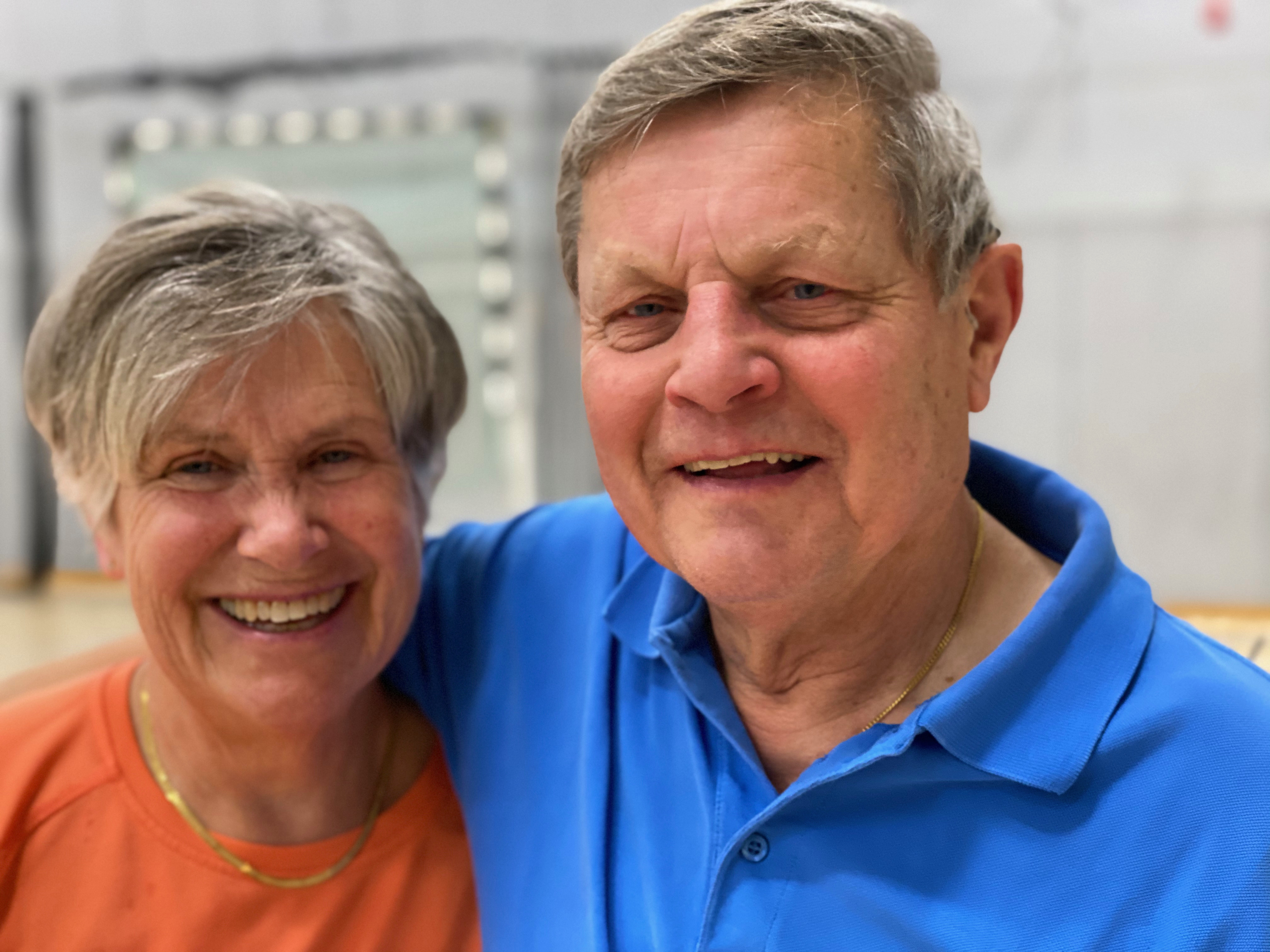 Ett äldre par står nära varandra och ler mot kameran i en inomhusmiljö.