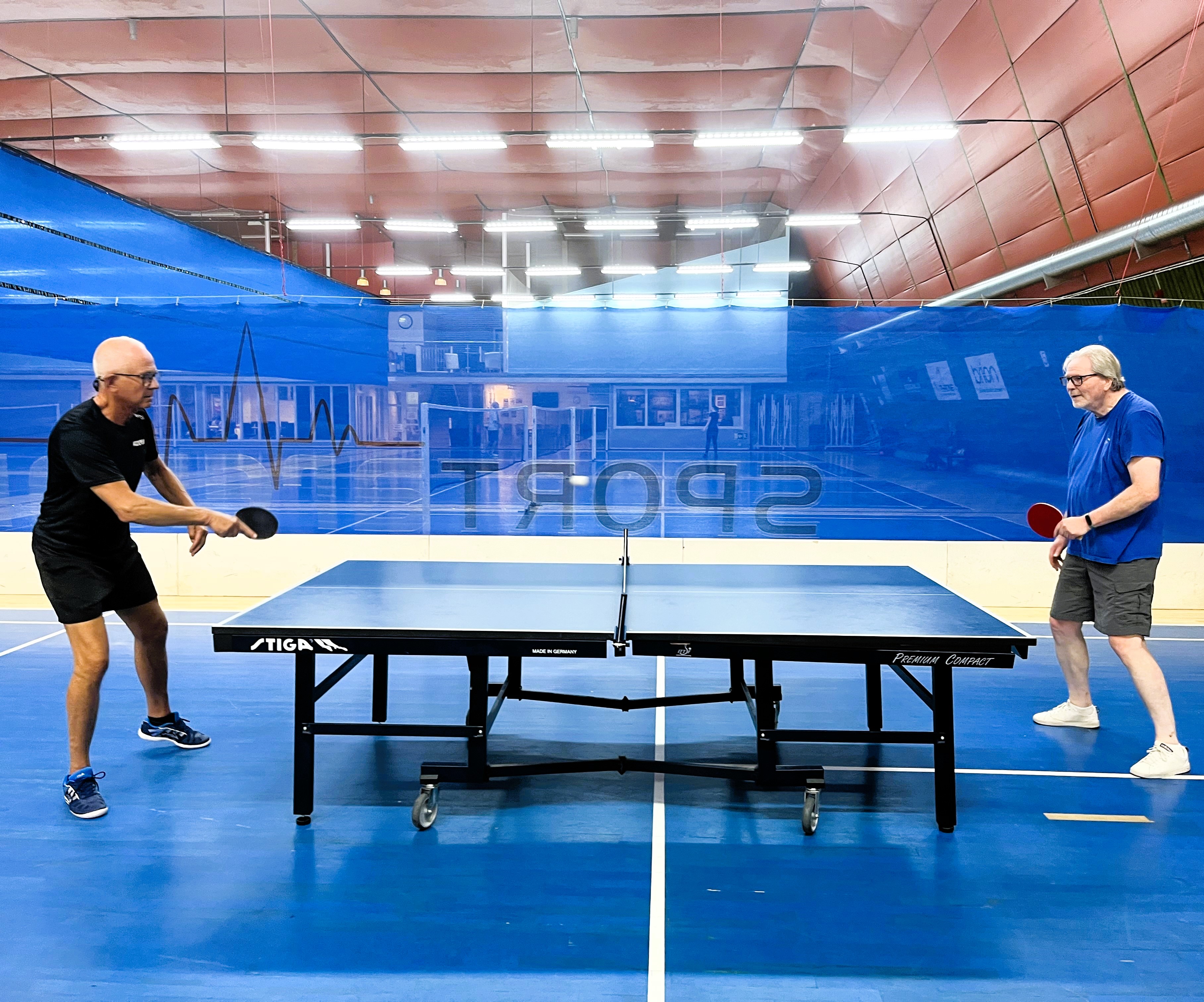 Två män spelar bordtennis mot varandra i en sporthall med blå väggar och röd takbelysning.