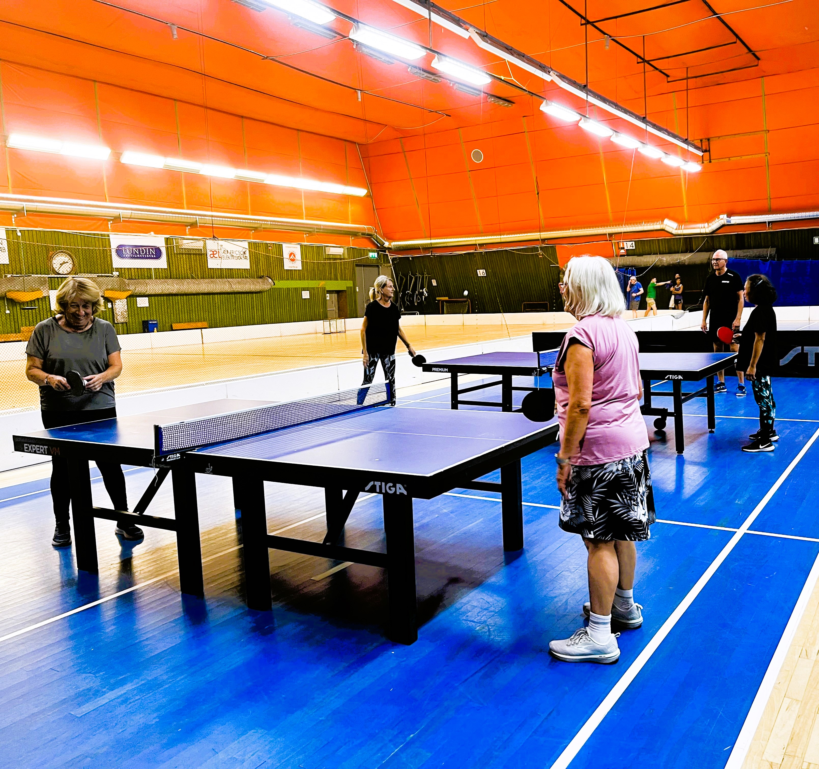 Flera personer spelar bordtennis i en stor hall med blått golv och orange tak.
