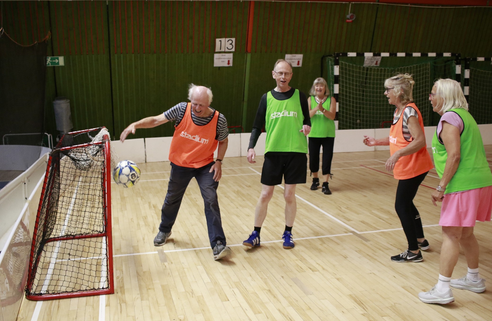 Äldre personer spelar gå-fotboll inomhus i gympasal. En spelare i orange väst gör mål i ett litet nät.