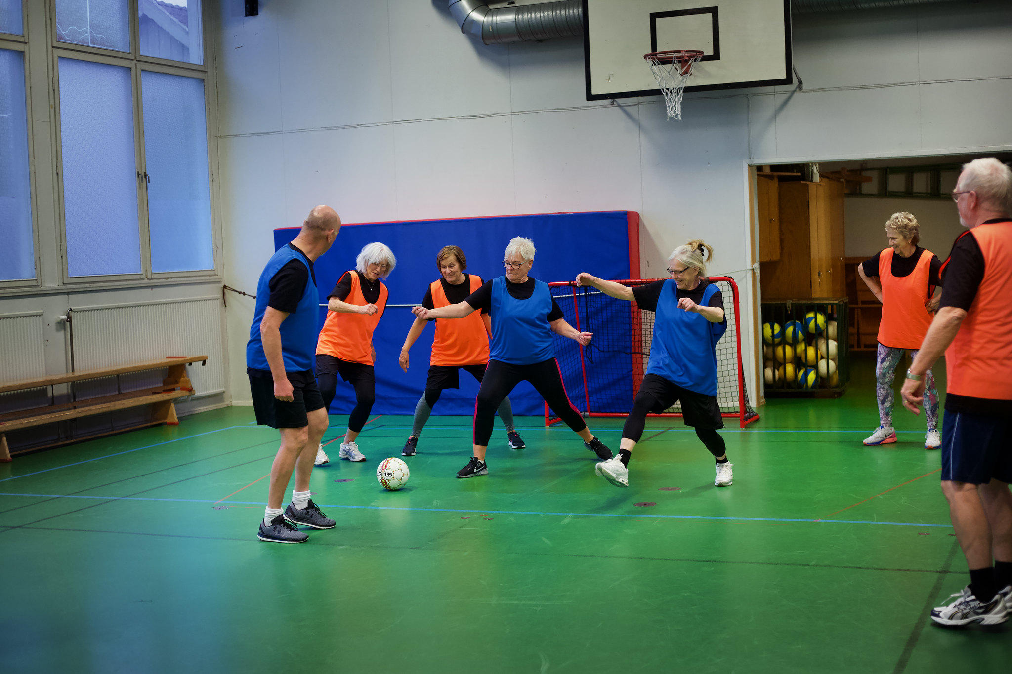 Äldre spelare i orange och blå västar spelar gå-fotboll inomhus. Flera deltar aktivt runt bollen.