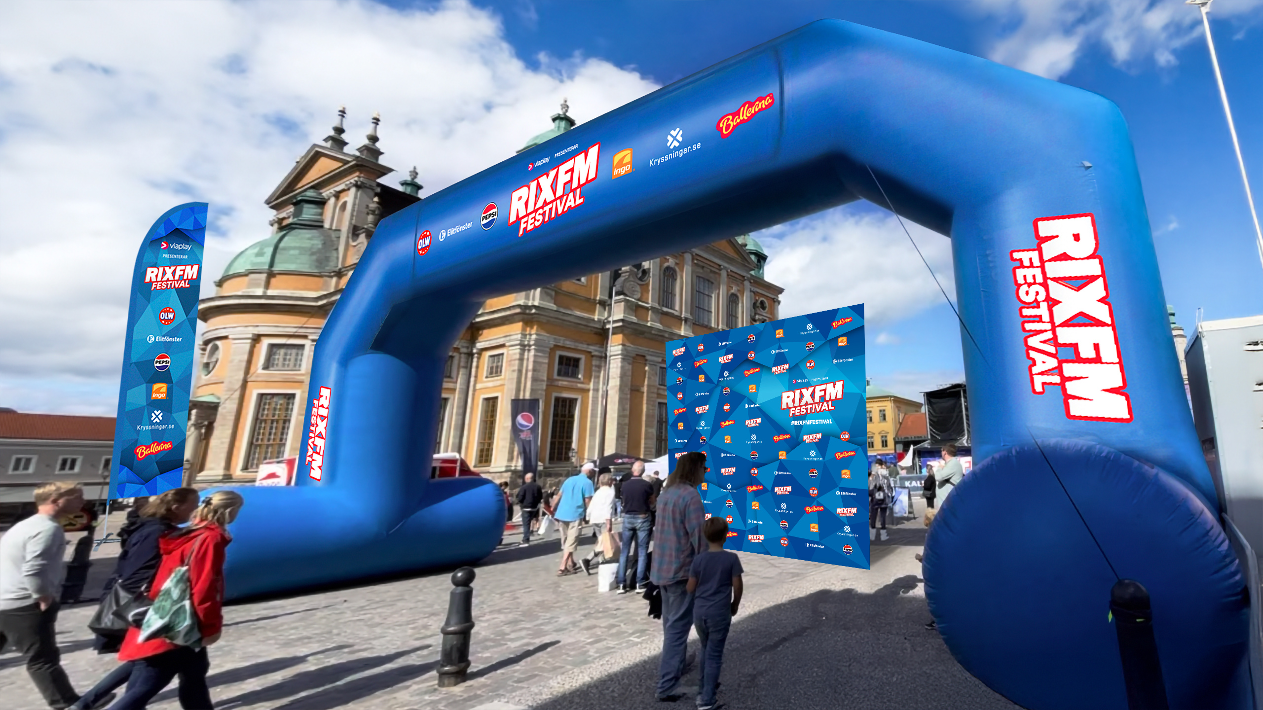 Ett stort blått uppblåsbart valv med texten RIX FM Festival står uppställt på ett torg framför en äldre kyrkobyggnad. Människor går in genom valvet mot ett område där en blå fotovägg med logotyper är uppställd.