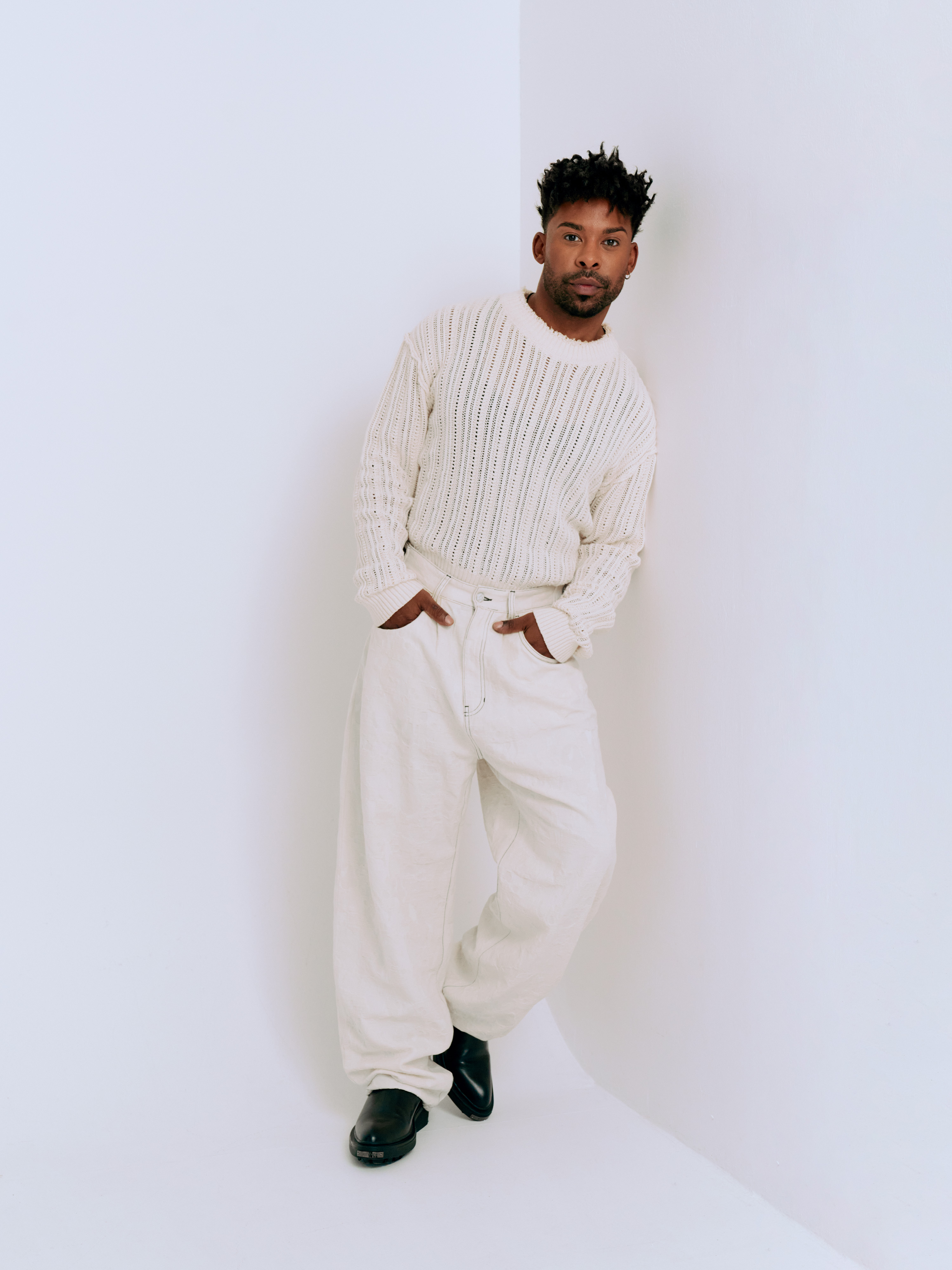 En man står lutad mot en vit vägg och tittar mot kameran. Han är klädd i en vit stickad tröja och vida vita byxor. Ena foten är lätt framför den andra.
