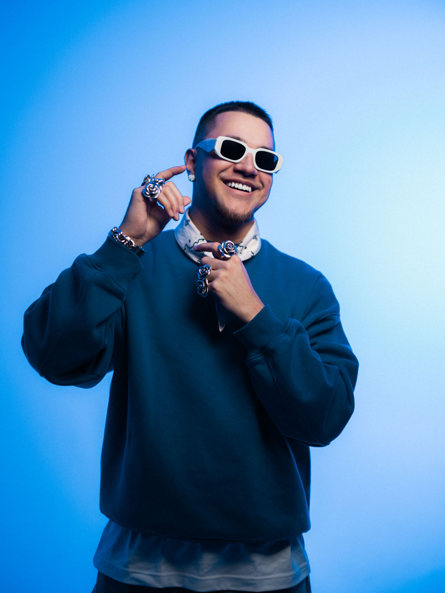 Artist Liamoo. En leende man med solglasögon och mörkblå tröja poserar mot en ljusblå bakgrund och håller i sitt halsband med båda händerna.