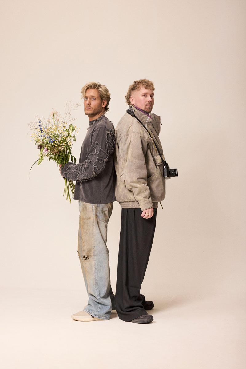 Två unga män står rygg mot rygg i en studio, en håller en bukett med vilda blommor. Båda bär lager-på-lager-kläder i streetwear-stil.
