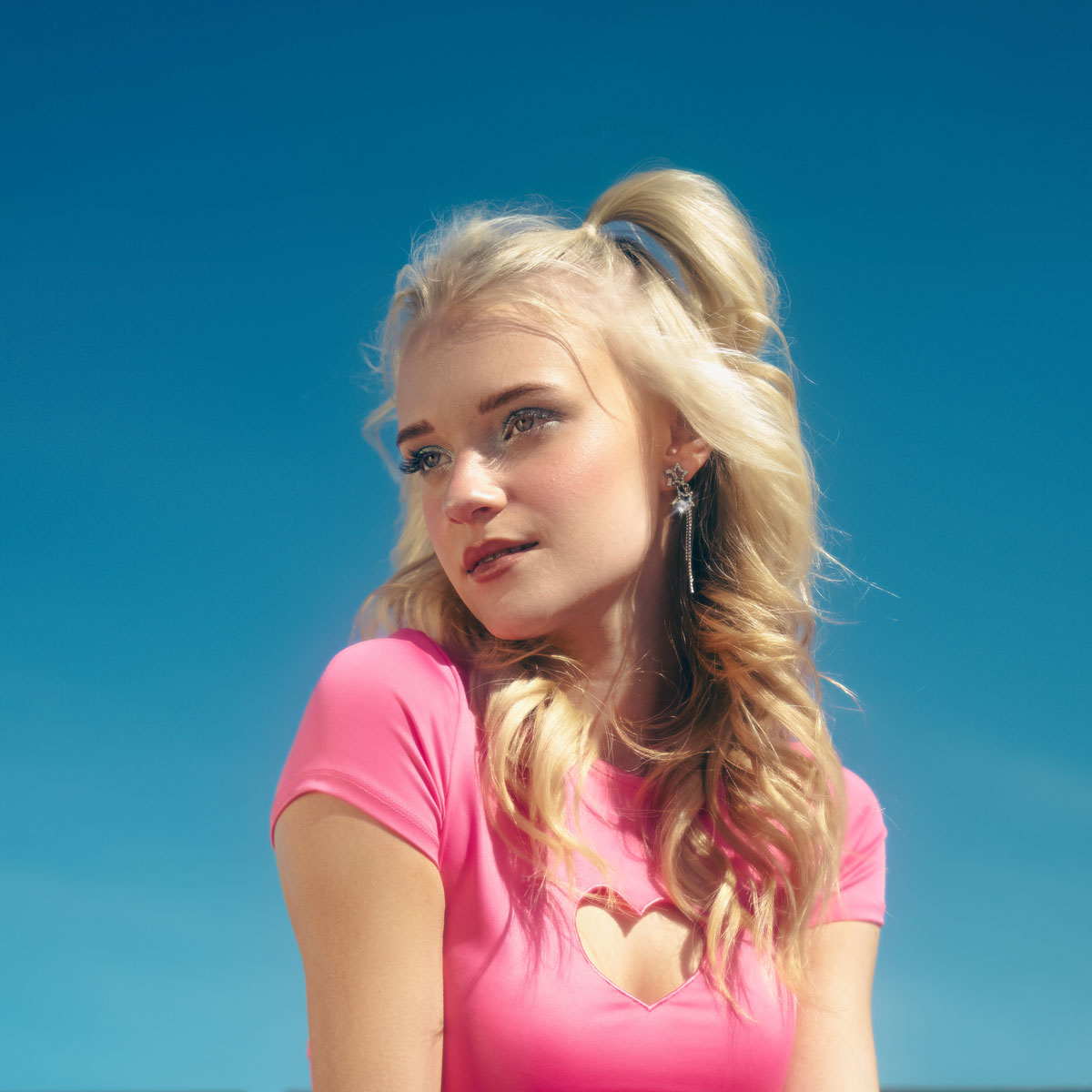 En blond kvinna i en klarrosa topp står mot en blå himmel och tittar lite åt sidan med ett mjukt leende.