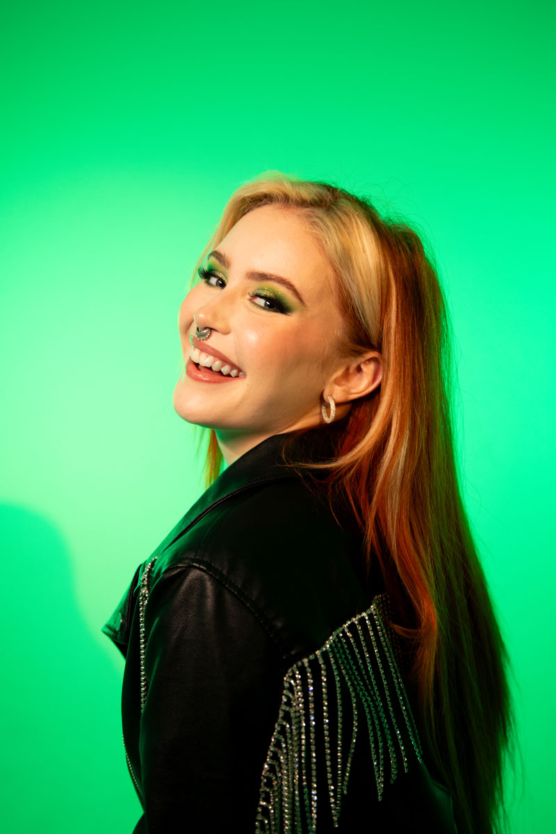 En leende kvinna med rödblont hår står vänd snett bort från kameran och tittar tillbaka över axeln. Bakgrunden lyser i grönt.