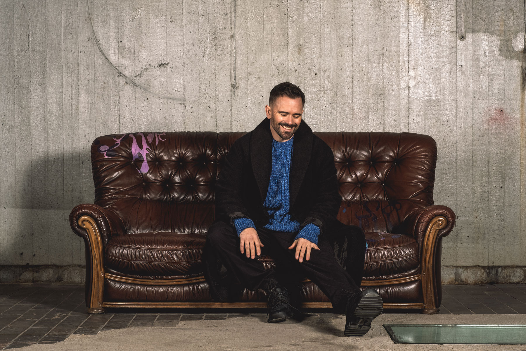 En man i svart och blått sitter skrattande på en sliten brun lädersoffa framför en betongvägg.
