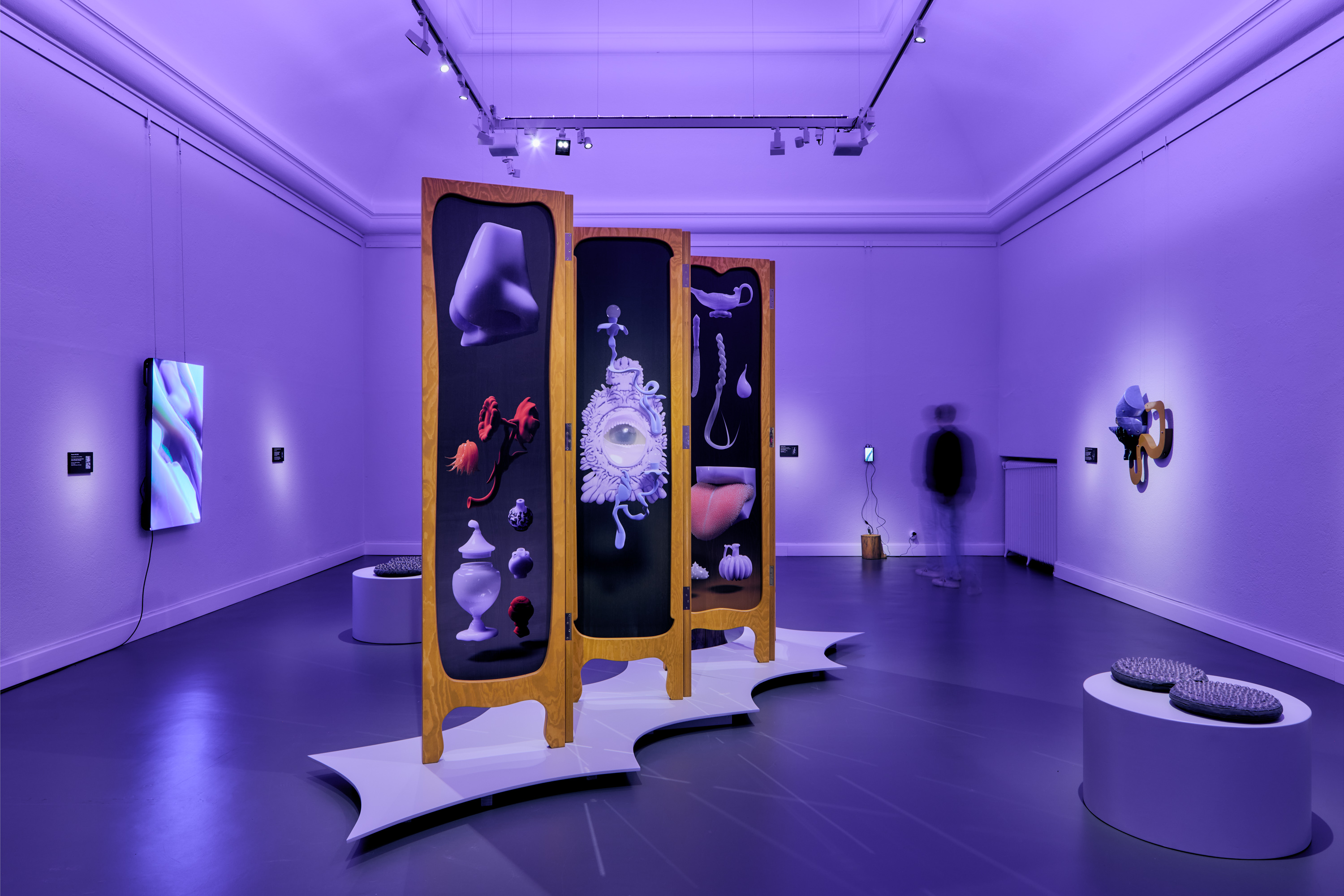 Ett museumrum i lila belysning med modern konst på väggarna och ett stort tredelat skärmverk i mitten med surrealistiska objekt.