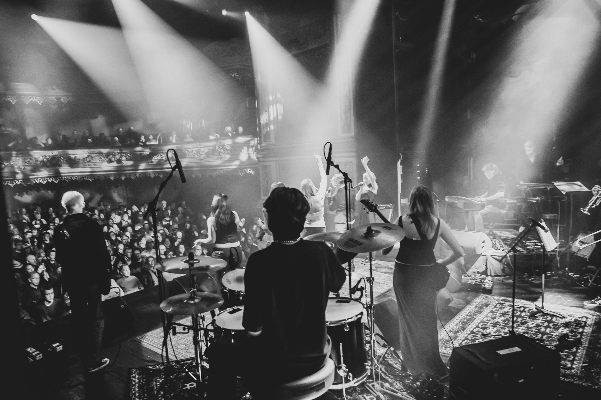 En svartvit bild visar ett liveuppträdande på en stor scen inför en entusiastisk publik. Bilden är tagen bakifrån scenen, där flera musiker syns spela – bland annat en trummis, en keyboardist och sångare.