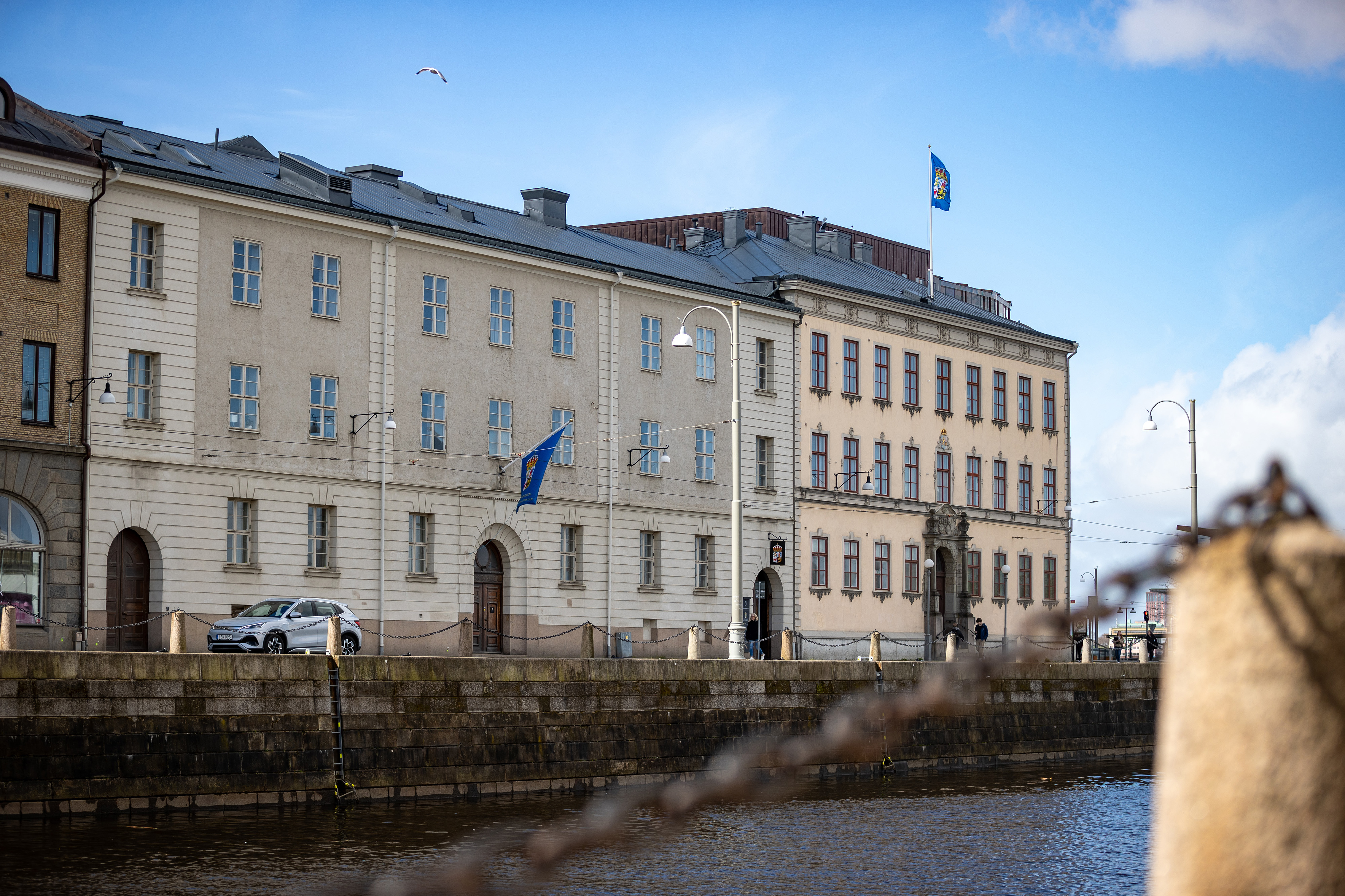 En ljus stenbyggnad med flaggor på fasaden och taket. Huset ligger vid vattnet under blå himmel. En kedja i förgrunden är suddigt synlig.