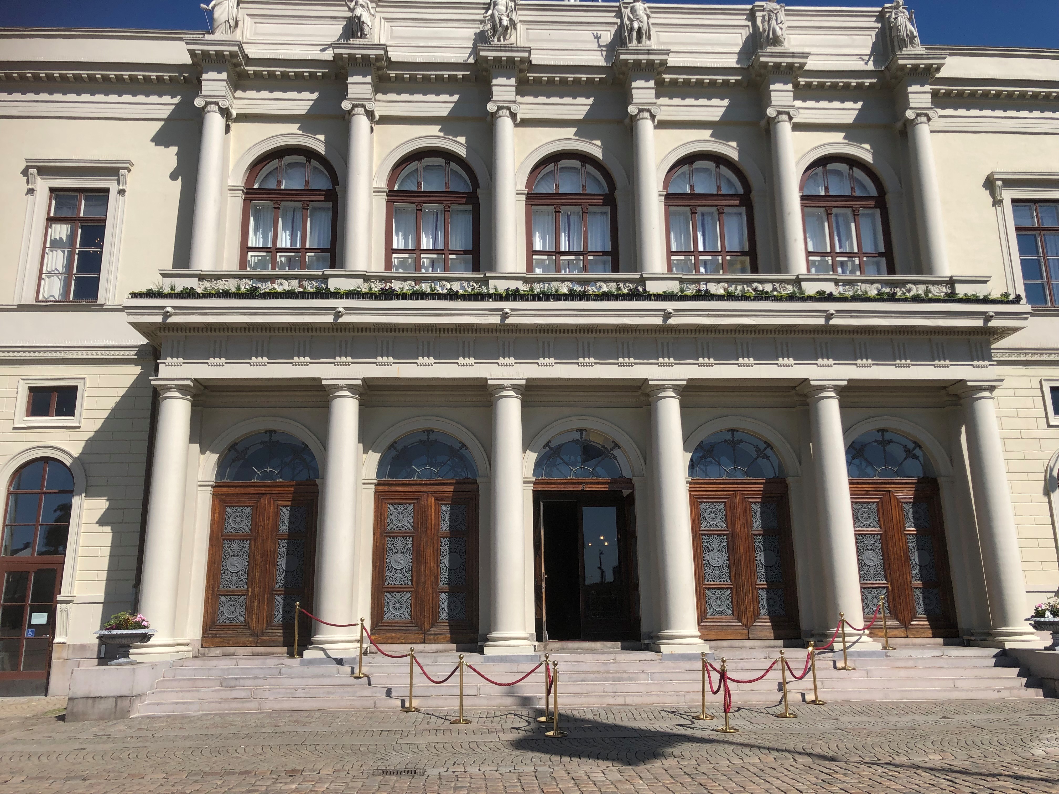 En pampig byggnadsfasad med höga pelare, stora välvda fönster och fem dubbeldörrar i trä. En av dörrarna står öppen. Framför entrén finns en stentrappa med röda rep som markerar ingången.