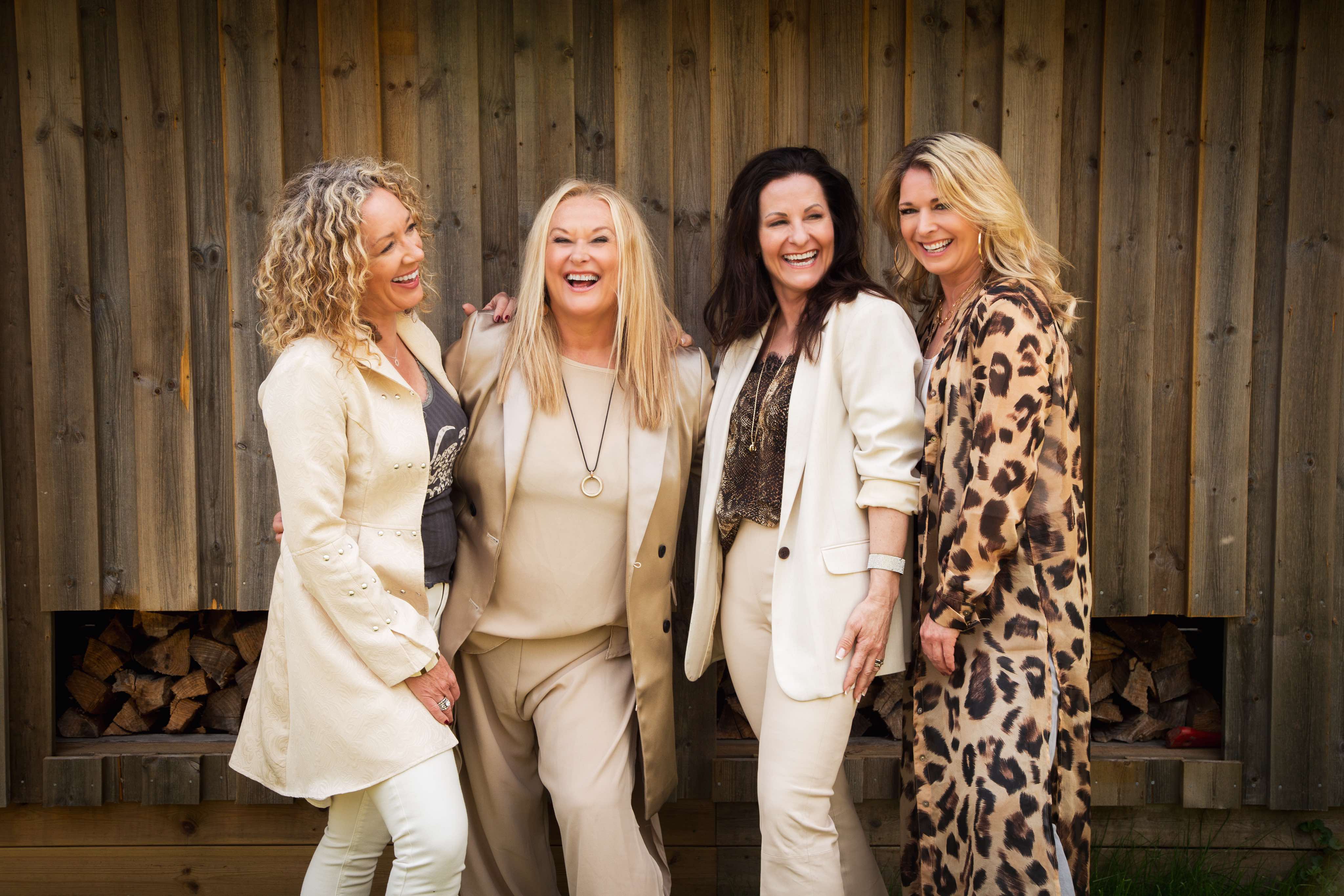 Fyra medelålderskvinnor i ljusa/beiga kläder ler och håller om varandra. I bakgrunden syns träpanel och ved staplat.