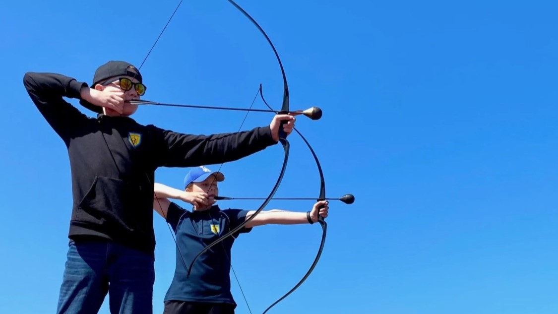 Två personer riktar varsin pilbåge mot måltavlor under klarblå himmel.