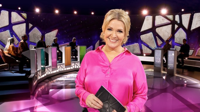 En kvinna med rosa skjorta/blus står i en tv-studio med talkort i händerna. Hon ler stort och i bakgrunden syns ljus, färger och personer som sitter vid varsin panel.
