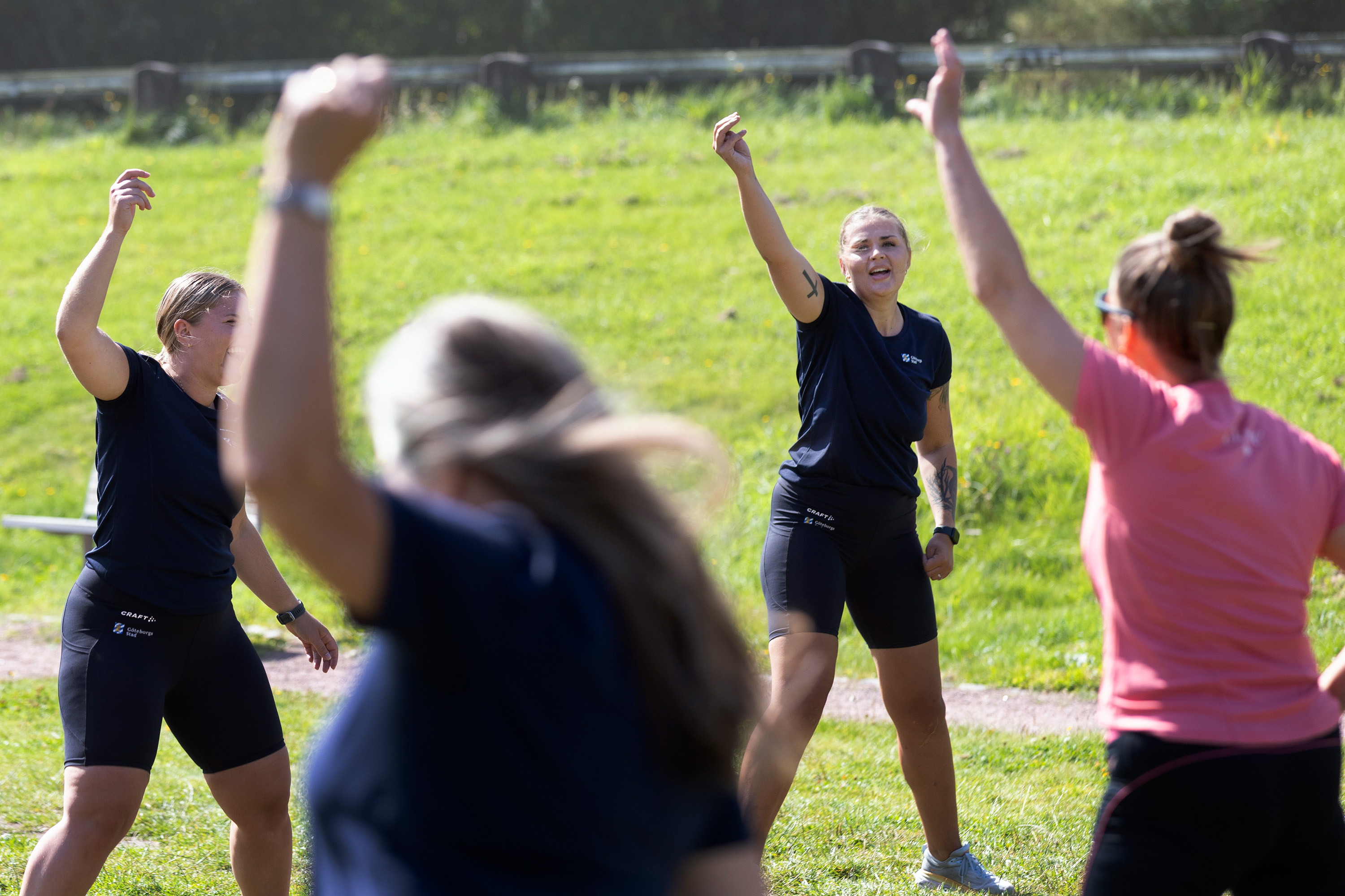En grupp vuxna personer tränar utomhus på en gräsyta. De står i ring och lyfter armarna uppåt i en gymparörelse. Flera av deltagarna ler, och stämningen ser energisk och glad ut.