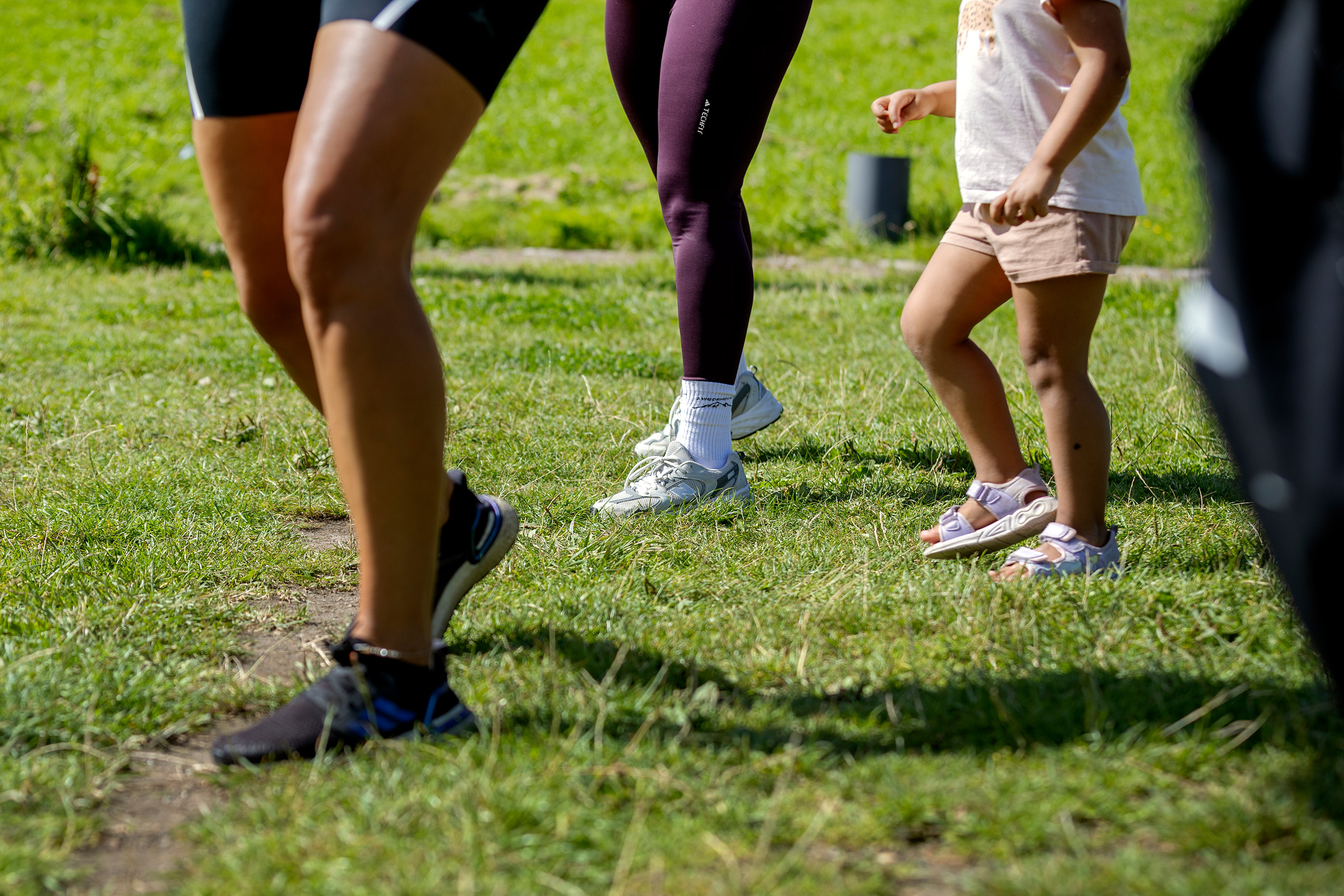 Närbild på ben och fötter av flera personer som tränar utomhus på gräs. En vuxen och ett barn deltar tillsammans, barnet har sandaler och shorts, medan de vuxna bär träningskläder och joggingskor.