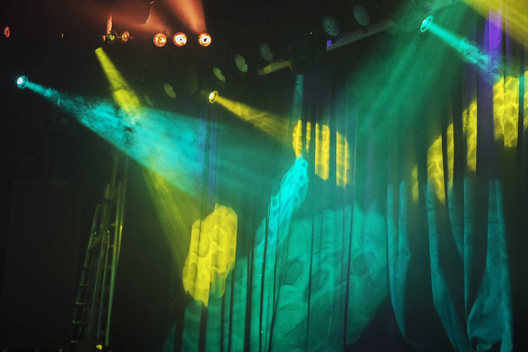 En scen belyst med färgade strålkastare i gult, blått och grönt. Ljuset skapar mönster på en neddragen ridå och scenutrustning syns delvis i bakgrunden.
