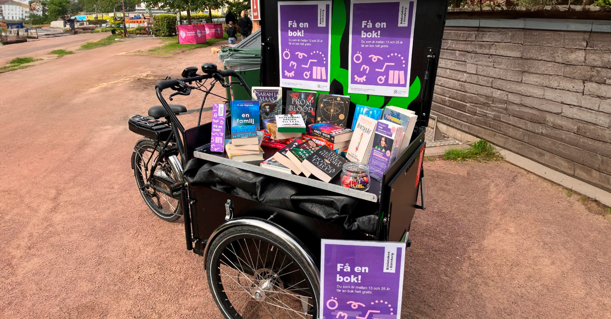 En lådcykel fylld med böcker står parkerad utomhus. På cykeln sitter skyltar med texten