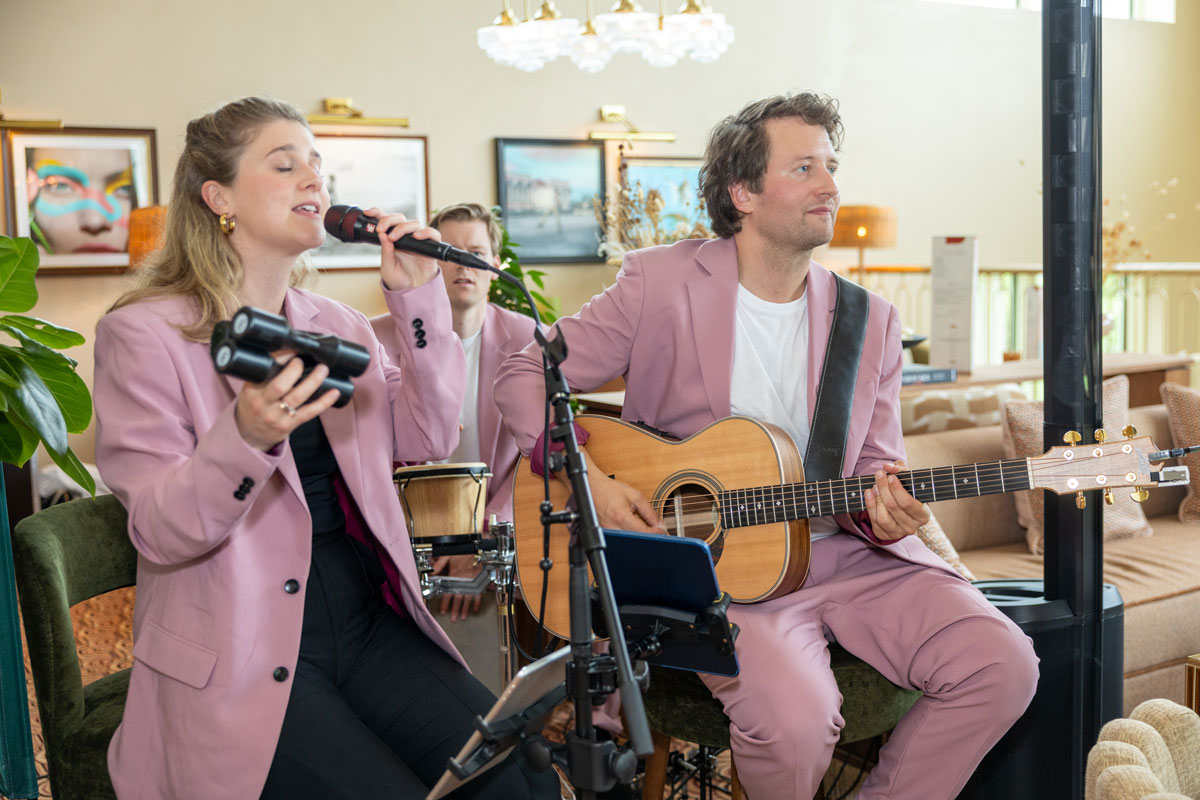 A woman and a man wearing matching pink blazers sit side by side. The man plays guitar while the woman holds a microphone and a shaker. They look happy.