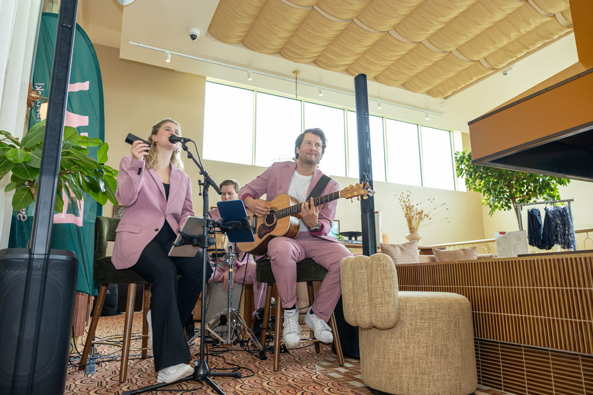 Hela musikgruppen syns i en öppen lokal. Kvinnan sjunger och mannen spelar gitarr. De sitter på höga stolar, och lokalen har stora fönster och mjuka färger.
