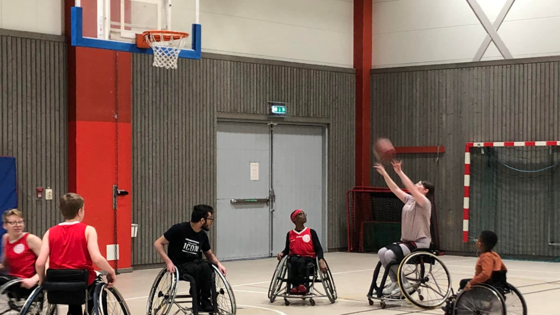 Sex personer spelar rullstolsbasket i en idrottshall. En spelare i ljus tröja är i luften med bollen och ser ut att skjuta mot korgen. Övriga spelare, varav flera bär röda tröjor, tittar mot bollen. I bakgrunden syns en grå vägg, en dörr och ett innebandymål.