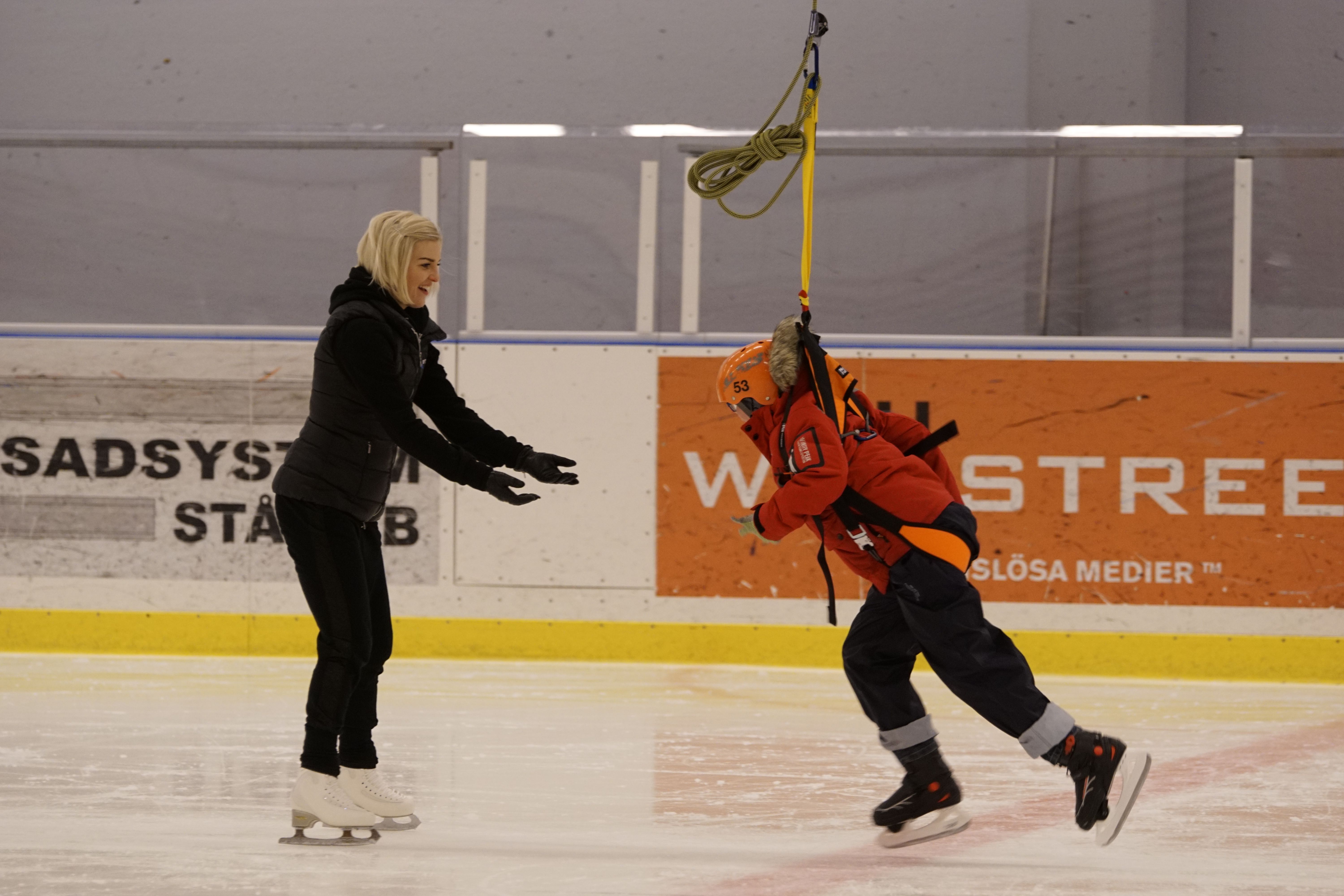Ett barn i hjälm och sele svävar ovanför isen i en ishall, fastspänt i en sele från taket. En vuxen på skridskor sträcker ut armarna mot barnet för att ta emot det.