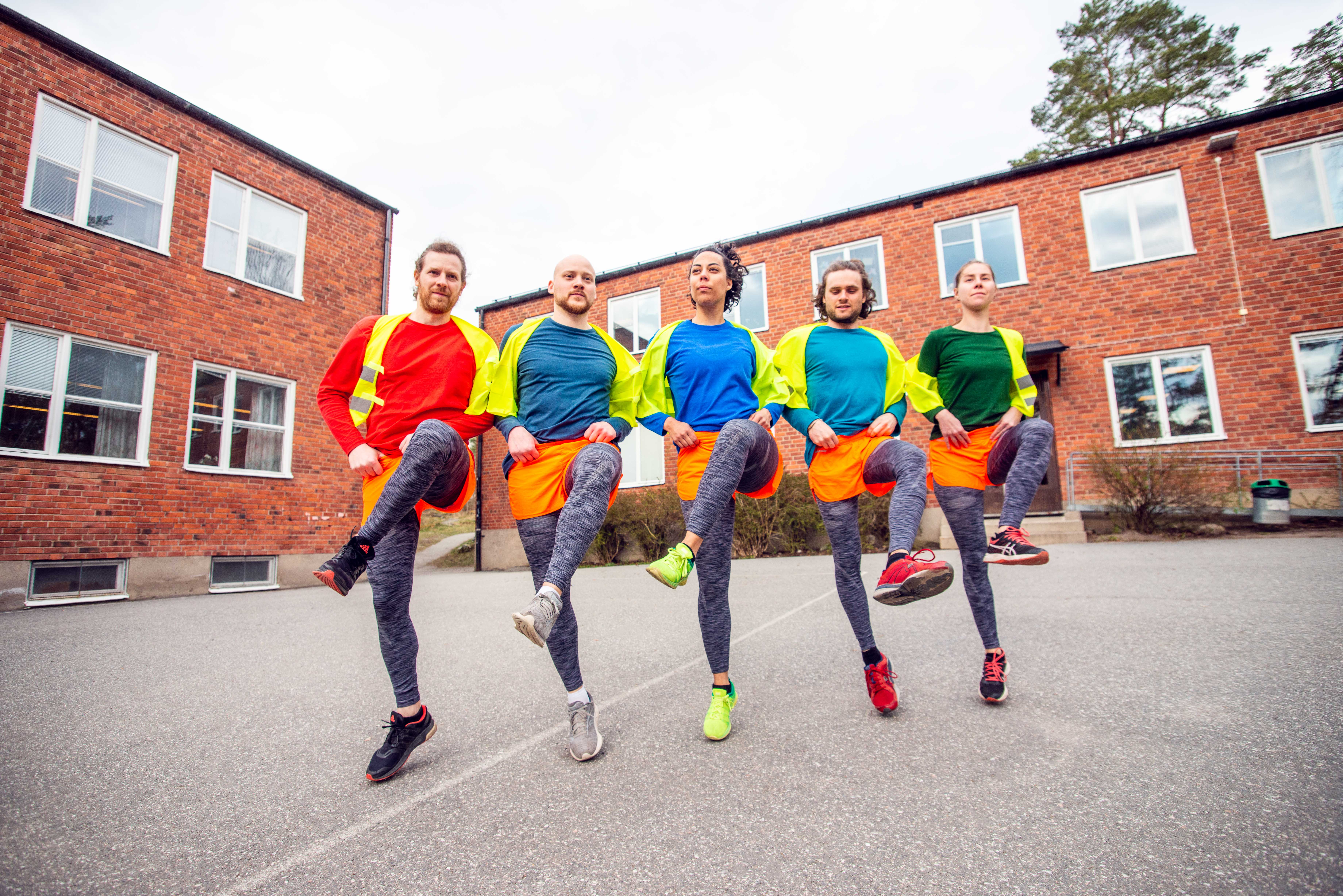 Fem personer står i rad på en asfalterad gård framför en röd tegelbyggnad. De har färgglada kläder i rött, blått, grönt och orange med reflexvästar och mörka tights. Alla lyfter sitt vänstra knä i luften i en synkroniserad rörelse, som i en dans eller uppvärmningsövning.