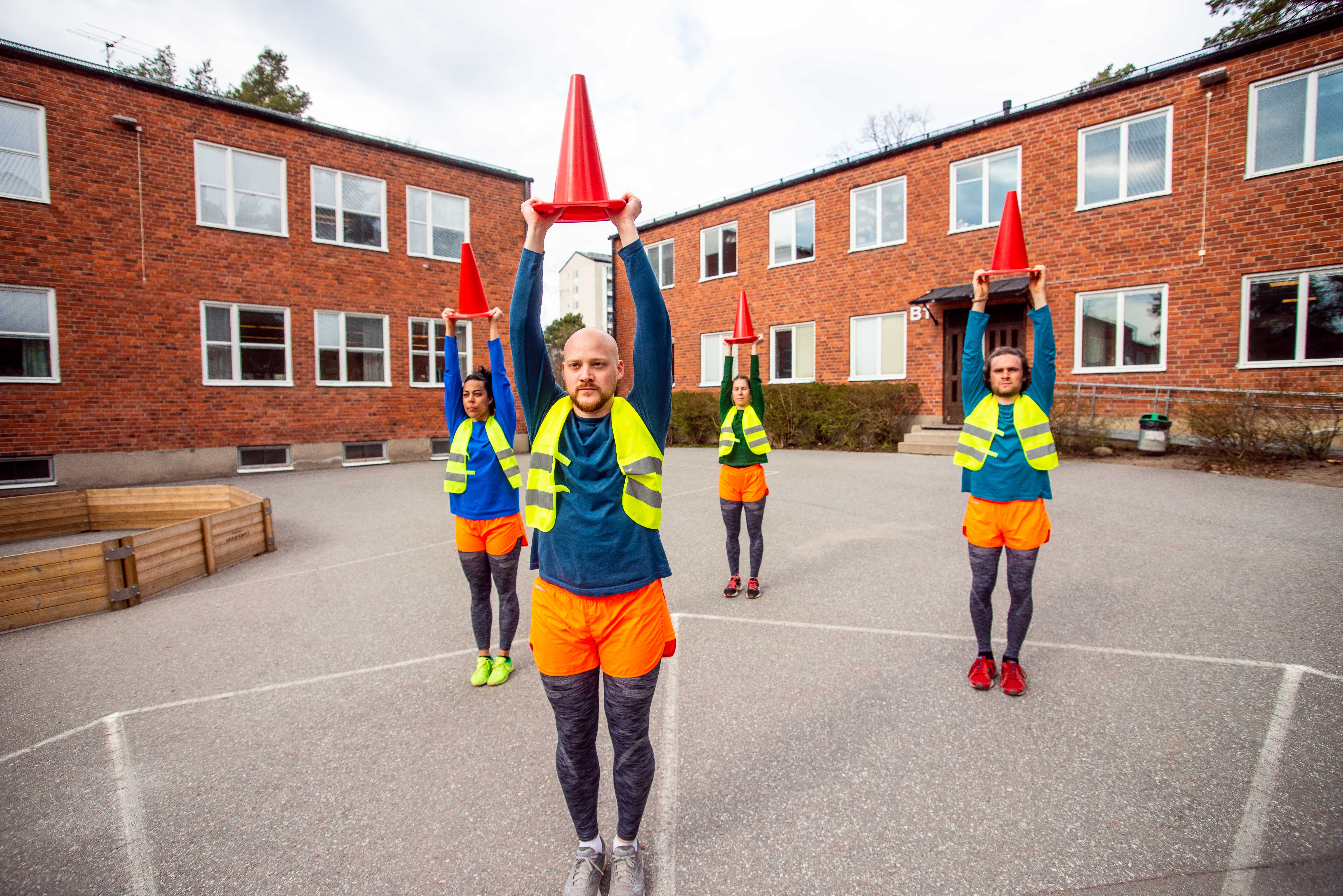 Fem personer står på en asfalterad gård framför en röd tegelbyggnad. De håller varsin stor trafik-kon ovanför huvudet. Alla har färgglada kläder och reflexvästar.