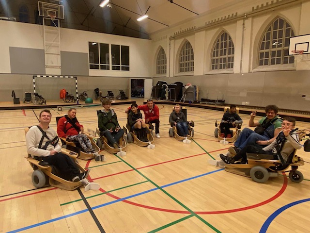 En grupp personer sitter i en gymnastiksal på specialbyggda, låga rullstolar med stora hjul. De sitter i en cirkel och ser glada ut – flera av dem vinkar eller gör tummen upp. Hallen är ljus med stora fönster och färgade linjer på golvet.